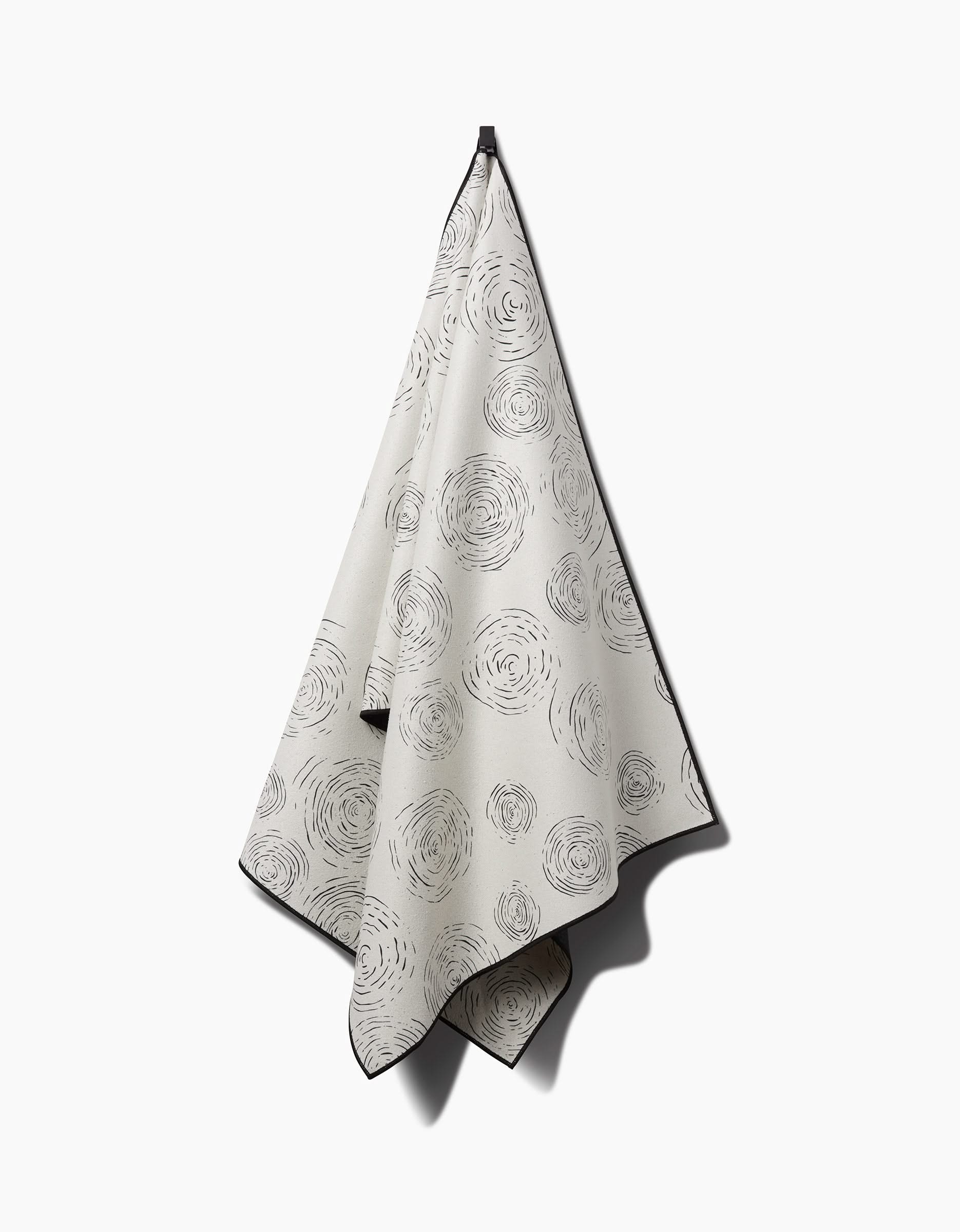 The GEOMETRY Rhythmic yoga towel, cream-colored with black, hand-drawn circular swirl patterns, hangs from a hook against a plain white background.