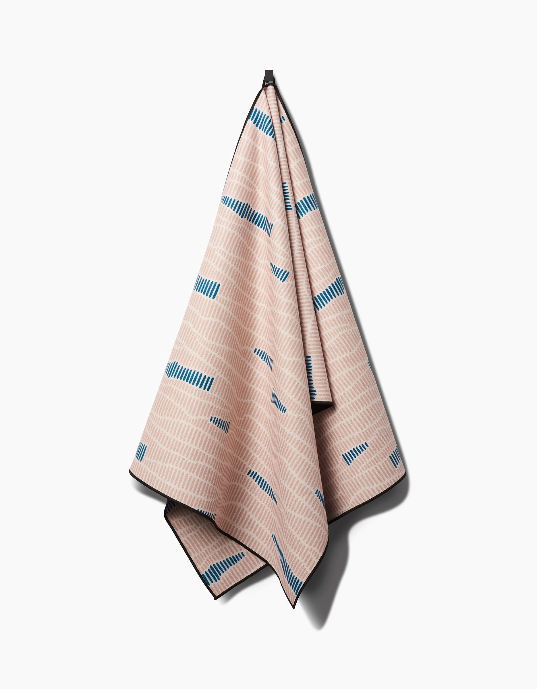 The Twig + Spark Ridgeline yoga towel, beige with a subtle zigzag pattern and blue rectangular accents, hangs from a single hook against a plain white background.