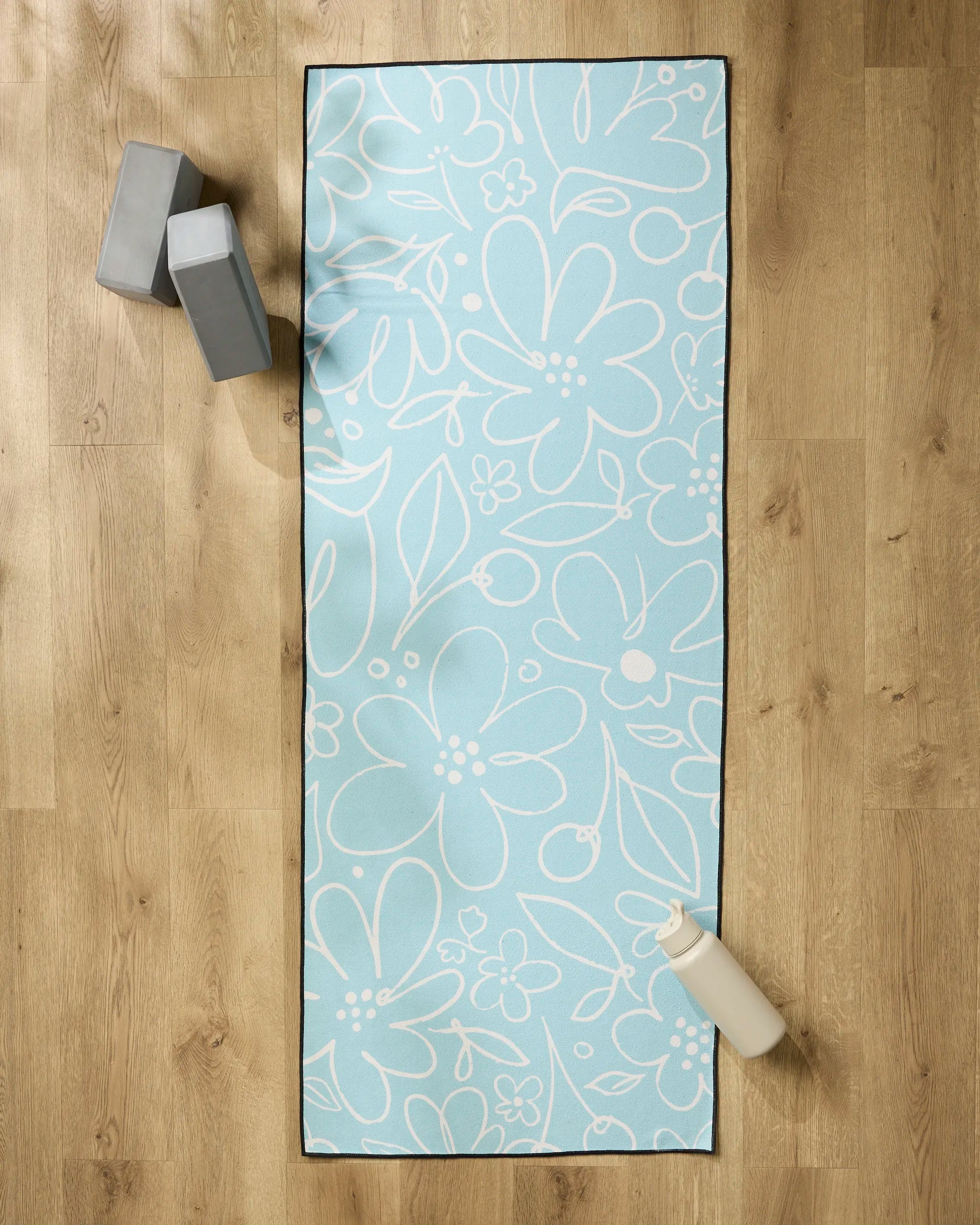 The Sara Mari River Blossom yoga mat, light blue with white floral patterns, lies on a wooden floor with a non-slip towel on top. Two gray blocks rest at the top left and a white water bottle sits at the bottom right corner.