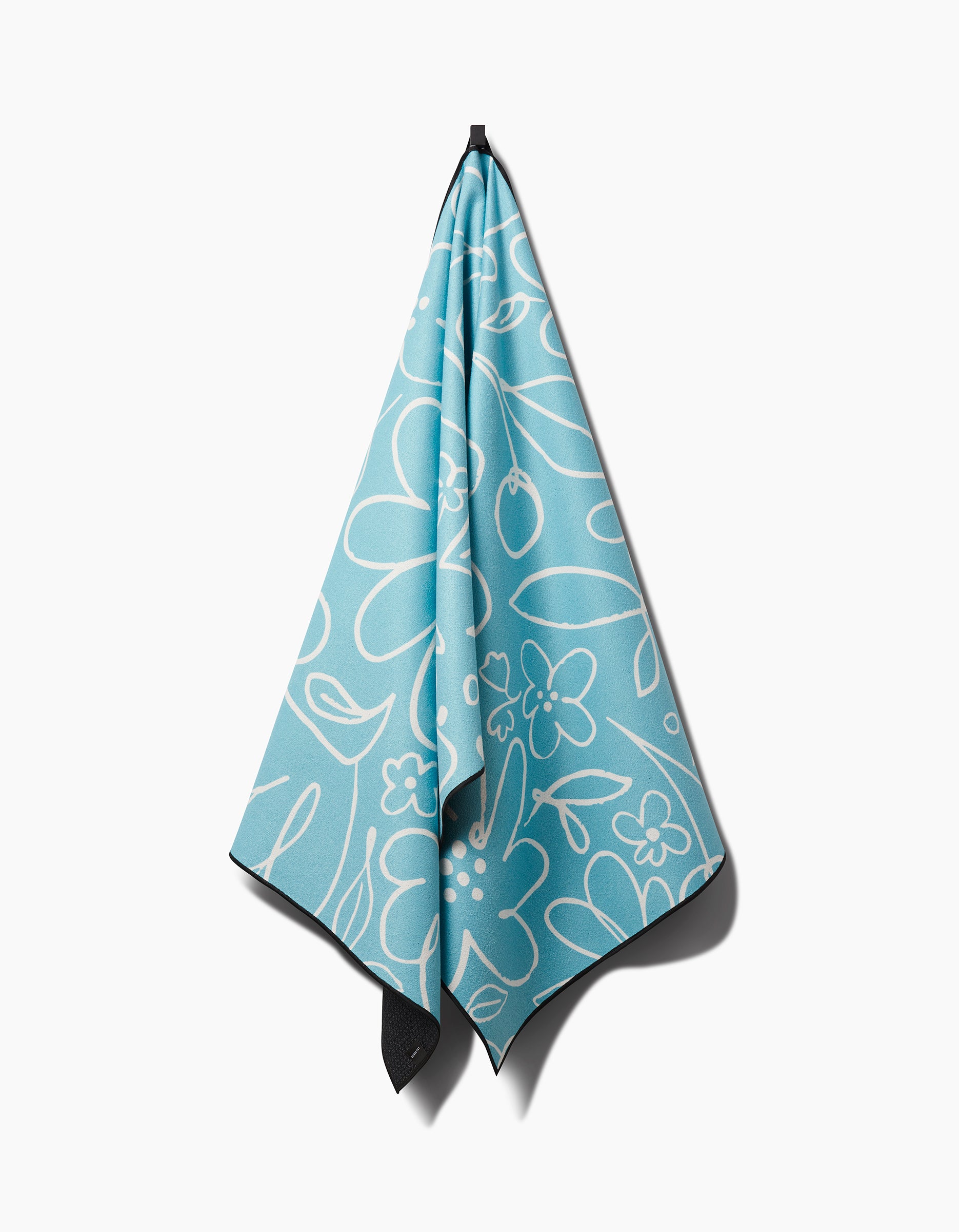 The Sara Mari River Blossom, a light blue yoga towel with a white floral print made from recycled materials, hangs on a black hook against a white background.