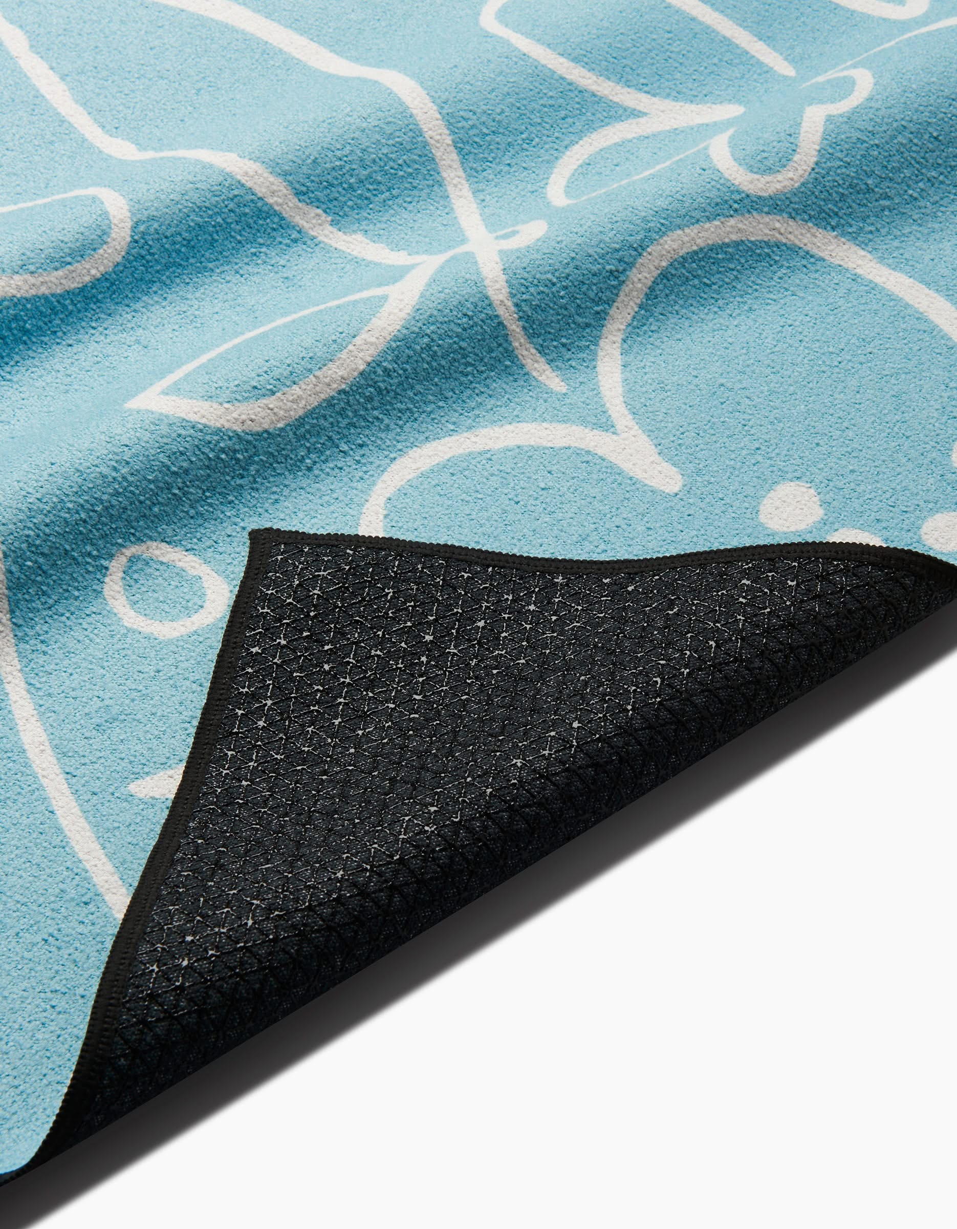 The Sara Mari River Blossom yoga towel features light blue fabric with abstract white lines and is partially folded to show its textured black non-slip backing made from recycled materials.