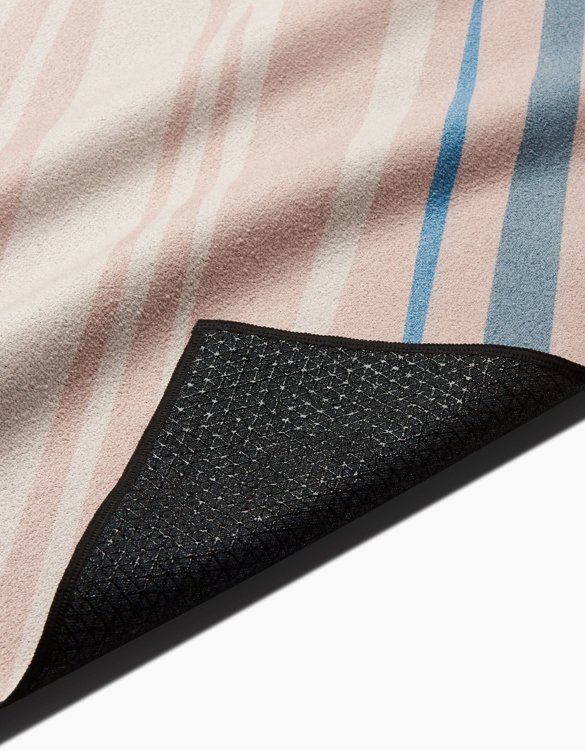 A close-up of the GEOMETRY Root yoga towel, rectangular and slip-resistant, features pastel pink, beige, and blue stripes with a black non-slip textured underside visible at one corner.