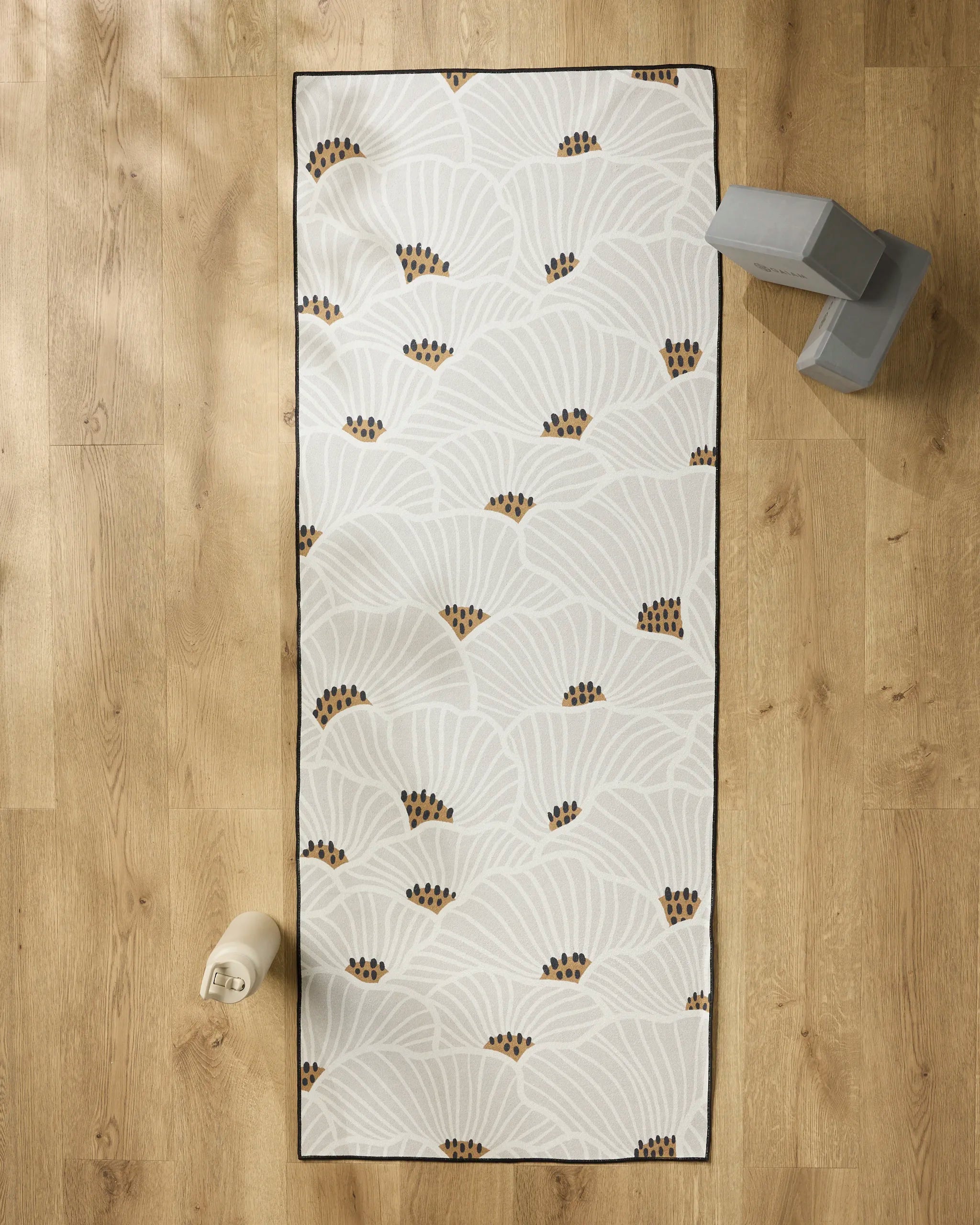 The Amy MacCready “San Diego” yoga mat with a fan-shaped, light-colored pattern and brown accents rests on a wooden floor, beside a white water bottle, gray yoga blocks, and a non-slip yoga towel.