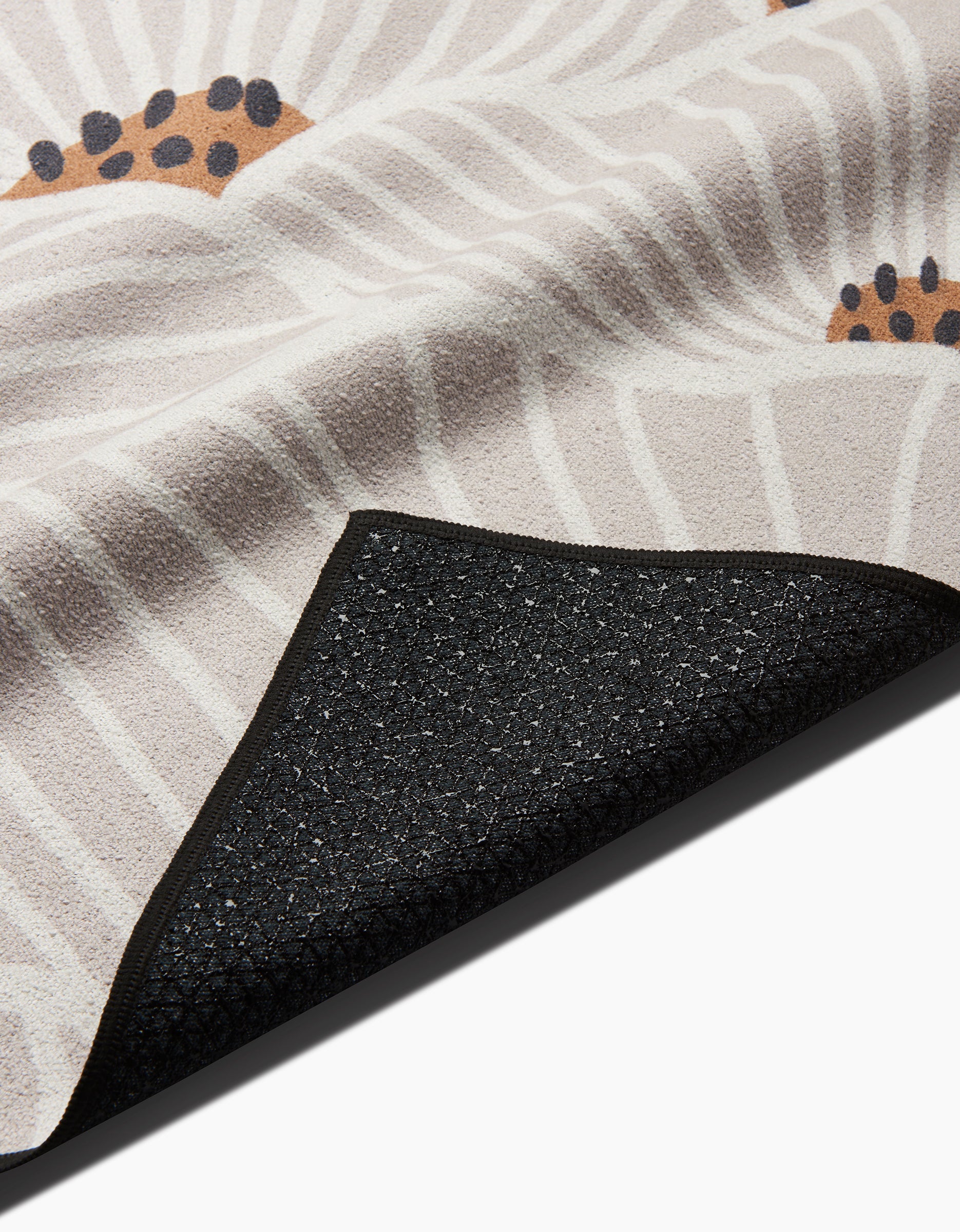 A close-up of the Amy MacCready San Diego rug features a beige, white, and black floral pattern. One corner is flipped to show its textured, slip-resistant black backing crafted from recycled materials.
