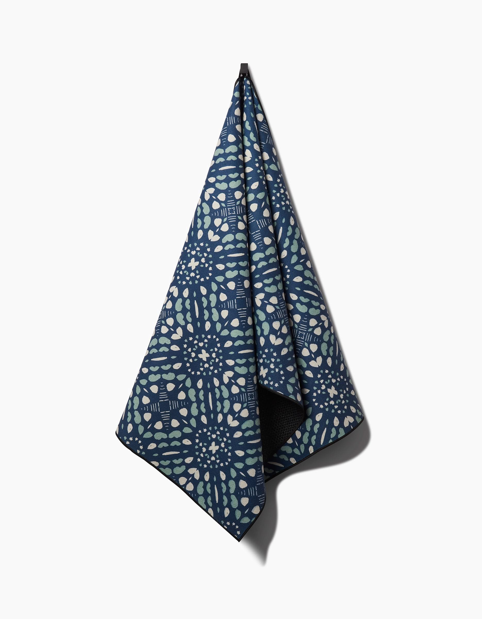 A Seeds and Nuts yoga towel by Twig + Spark, dark blue with a geometric pattern in light blue, green, and white shapes, hangs from a hook against a plain white background.