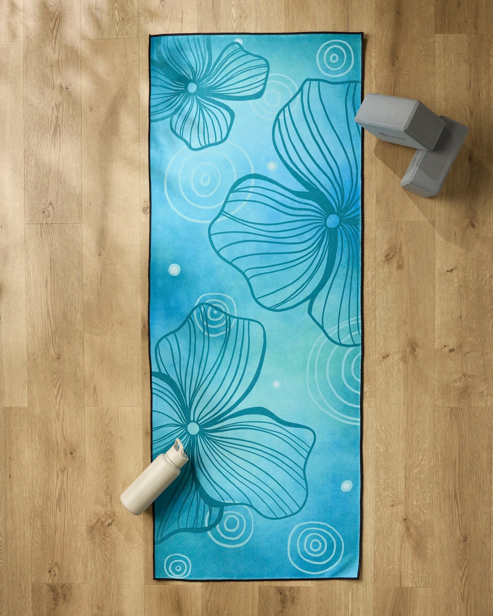 The Blissful Flamingo Serenity yoga mat, featuring abstract flower and swirl patterns, rests on a wooden floor with a white water bottle, two gray yoga blocks, and a non-slip towel for comfort and stability during practice.