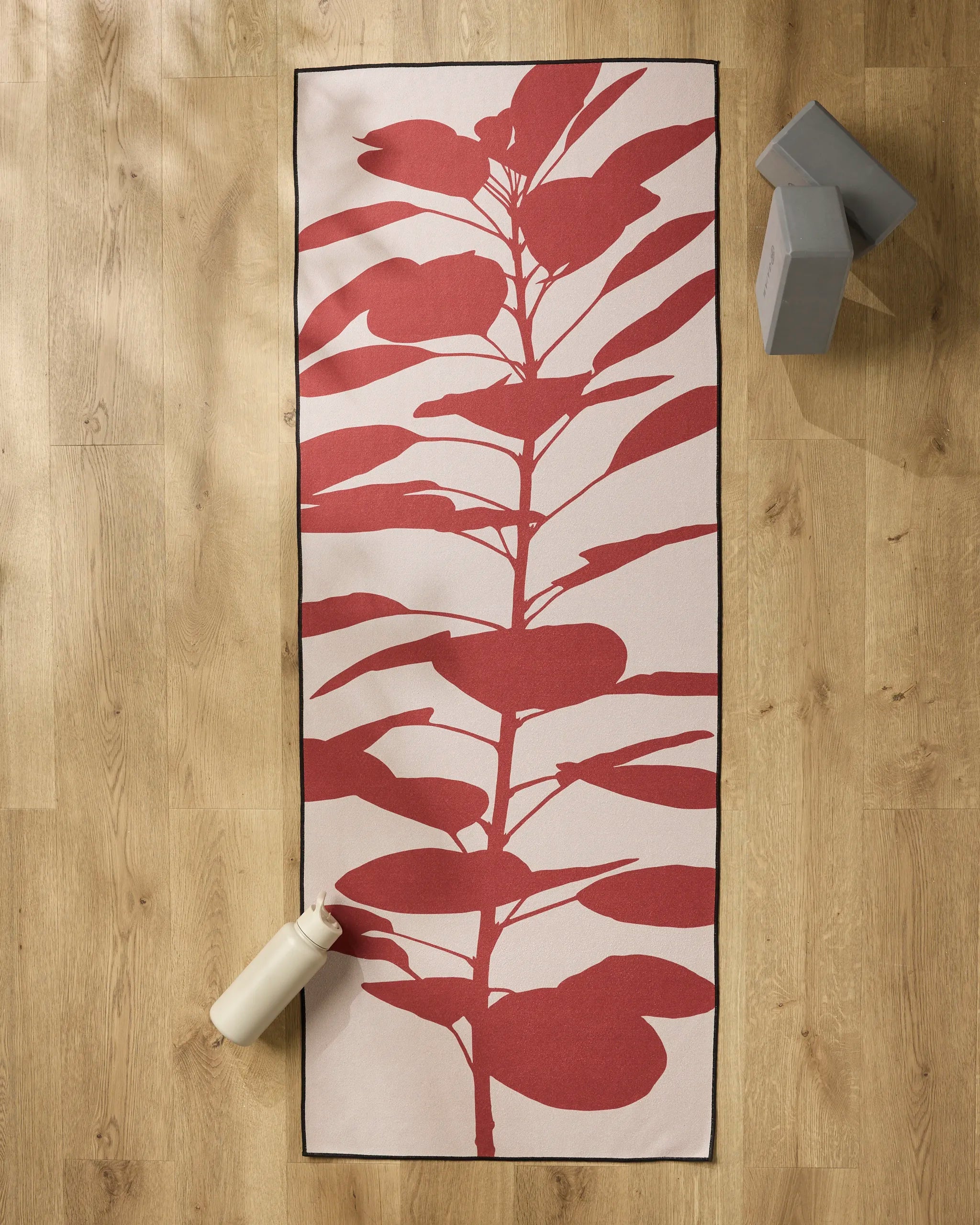 PrintedPlant's Smoketree yoga mat, featuring a large red leafy design, is placed on a light wooden floor with a white water bottle, two gray yoga blocks, and a slip-resistant towel for extra comfort during your practice.