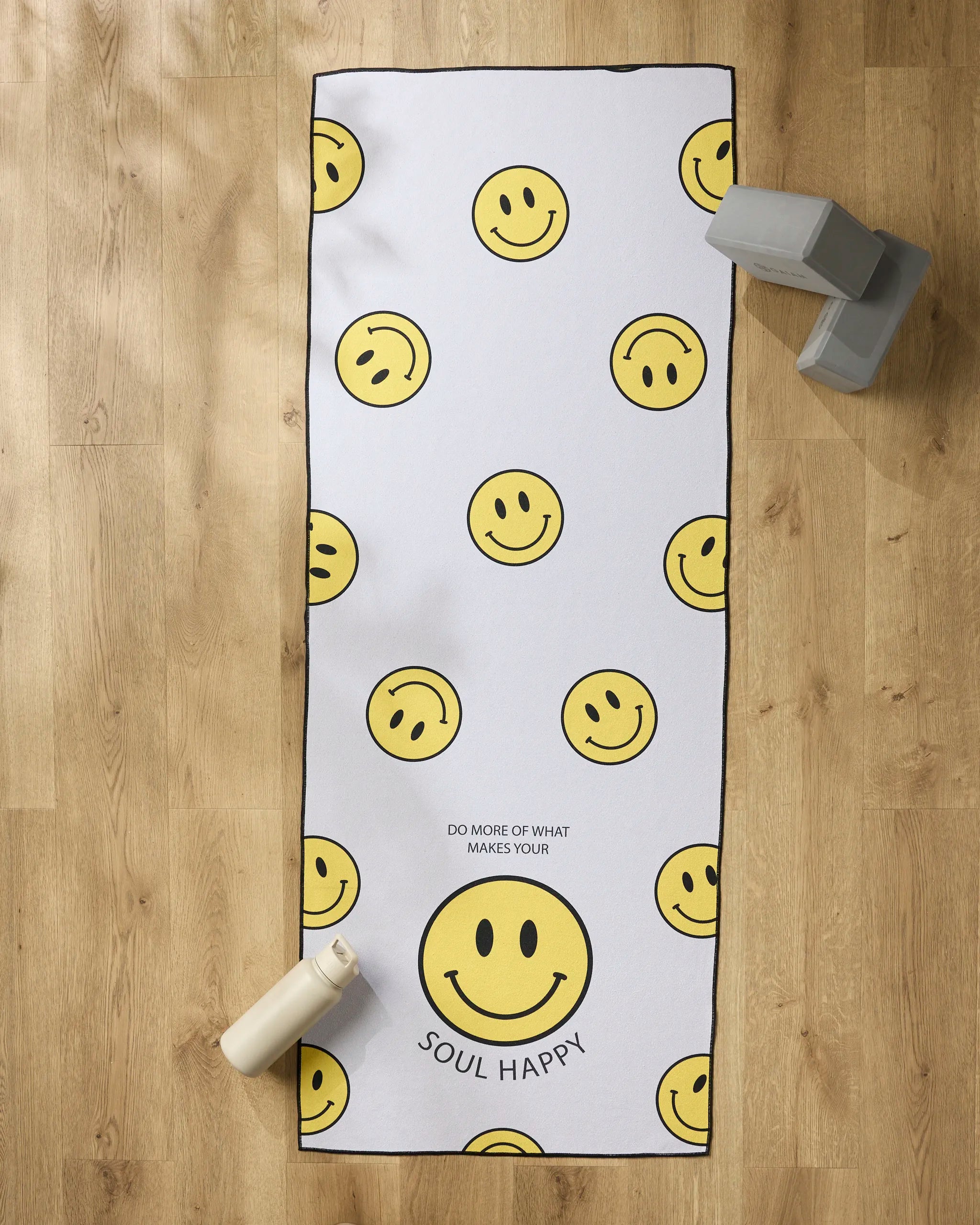 The Soul Happy yoga mat by Quotes by Christie features yellow smiley faces and "DO MORE OF WHAT MAKES YOUR SOUL HAPPY." Shown on a wooden floor with a non-slip towel, white water bottle, and gray yoga blocks for your practice.