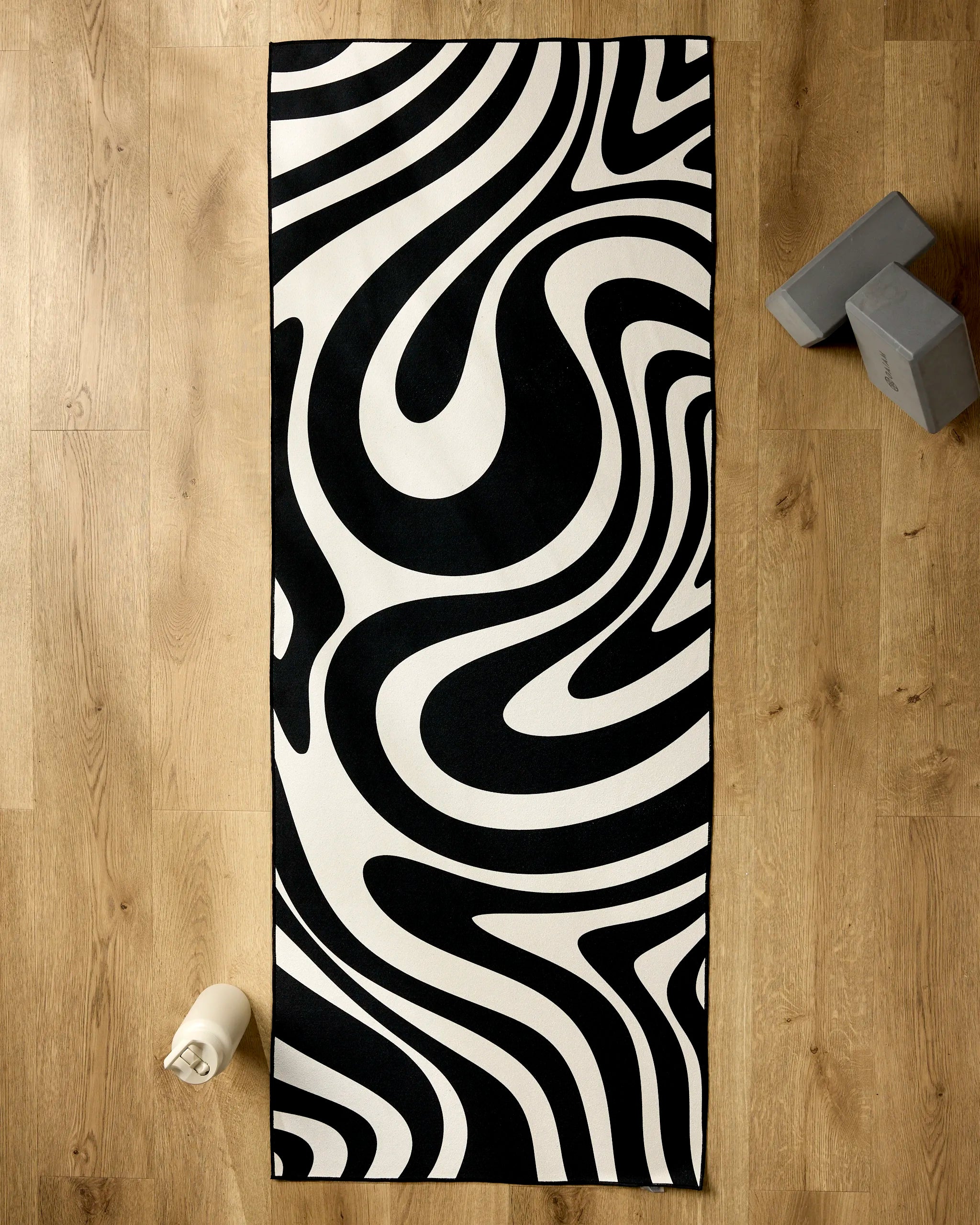 The GEOMETRY Spiral yoga mat features bold black and white abstract waves, shown on a light wooden floor with a recycled yoga towel. A water bottle and two gray blocks complete the setup.