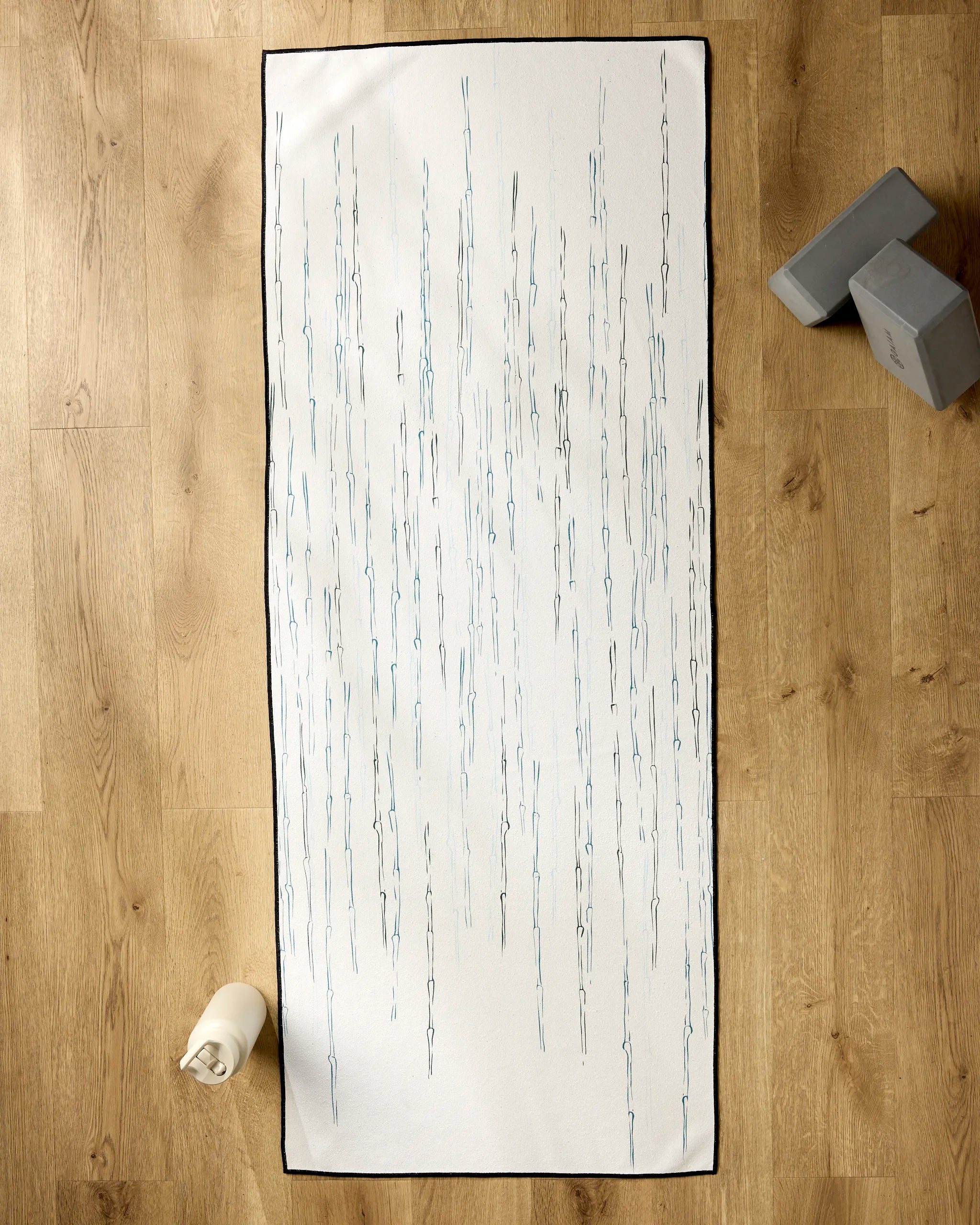 The GEOMETRY Stilts yoga mat, featuring a white background with vertical blue and gray lines, lies on a wooden floor alongside a rolled silicone grip towel and two gray yoga blocks.