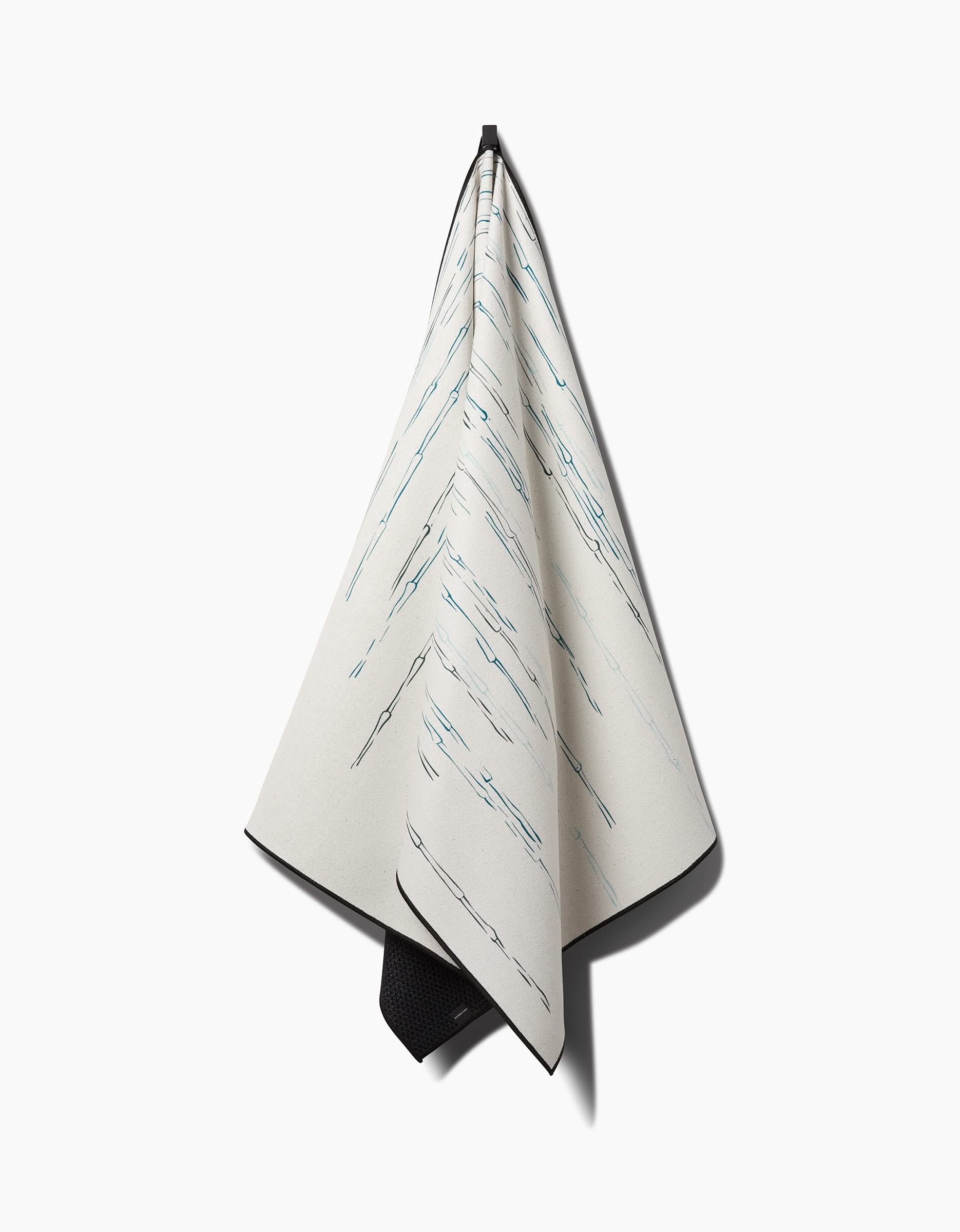 The GEOMETRY Stilts towel, featuring thin diagonal blue lines and a light, non-slip surface, hangs from a hook against a white background. Its simple, minimalistic design gives it a modern, elegant appearance.