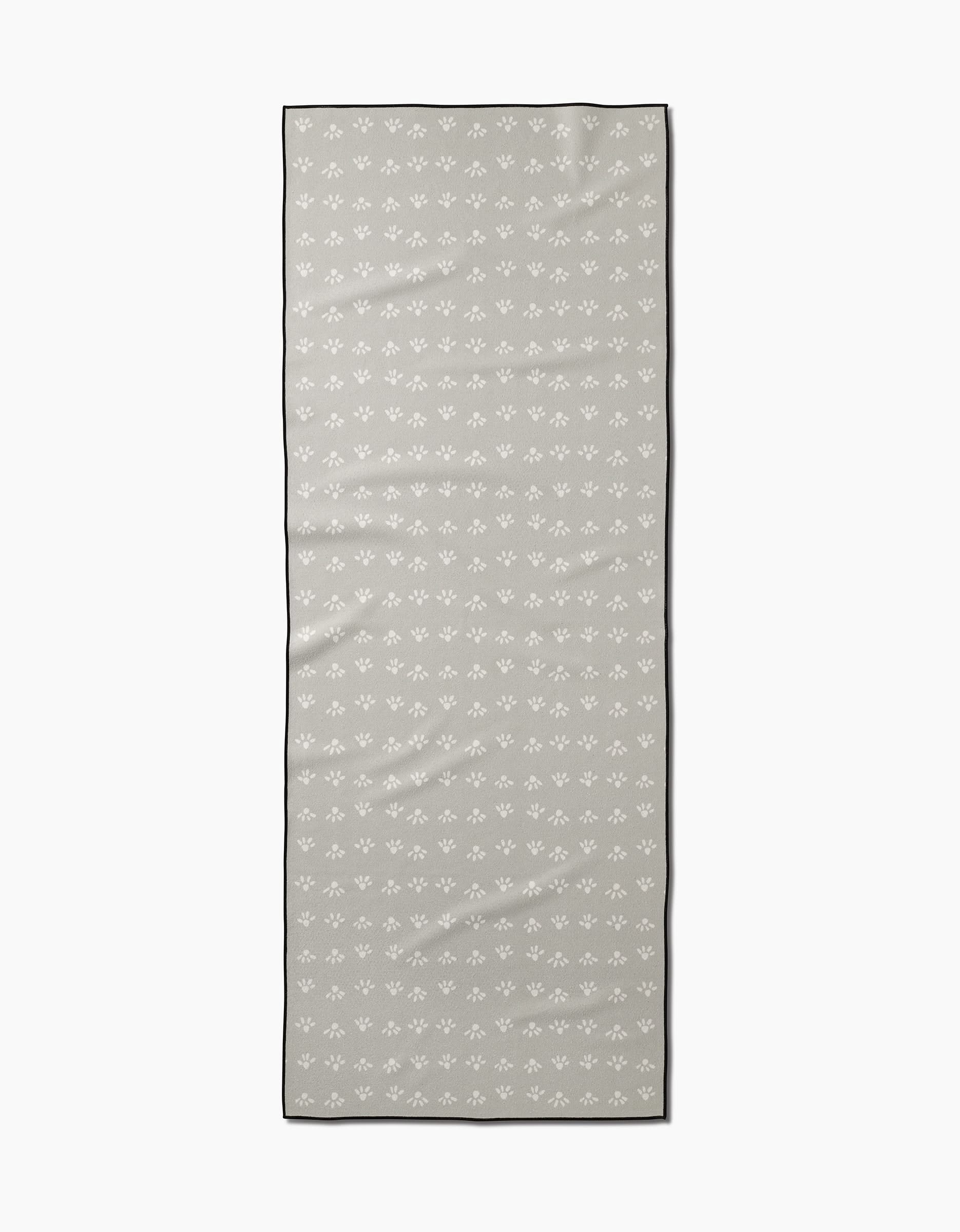 The Sun Bursts yoga towel by Anee Shah is rectangular, light gray, with a thin black border and a subtle pattern of small white fleur-de-lis motifs. It features a non-slip design and is shown on a plain white background.