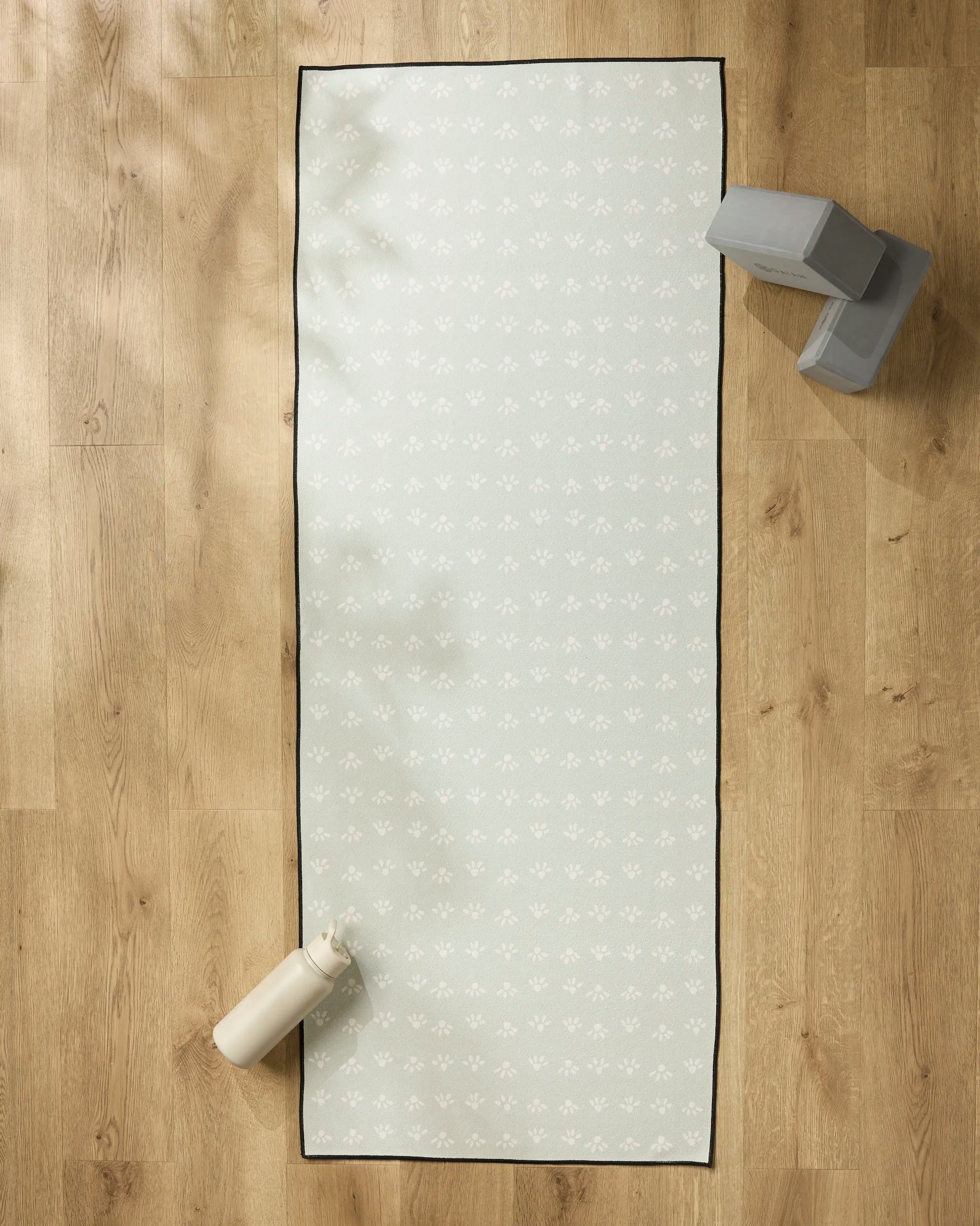 The Anee Shah Sun Bursts recycled yoga towel, featuring a subtle light pattern, is laid out on a wooden floor alongside a water bottle and two stacked yoga blocks. Sunlight casts gentle shadows across the serene scene.