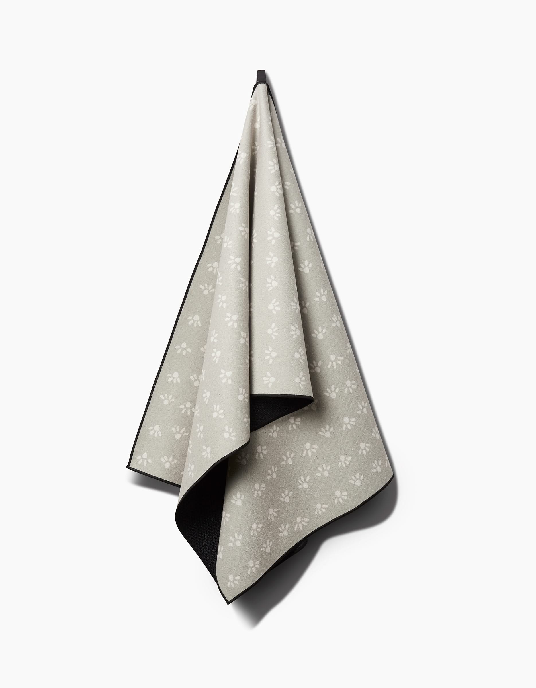 Anee Shahs Sun Bursts yoga towel, made from recycled material in beige and featuring white paw prints, hangs on a hook against a plain white background.