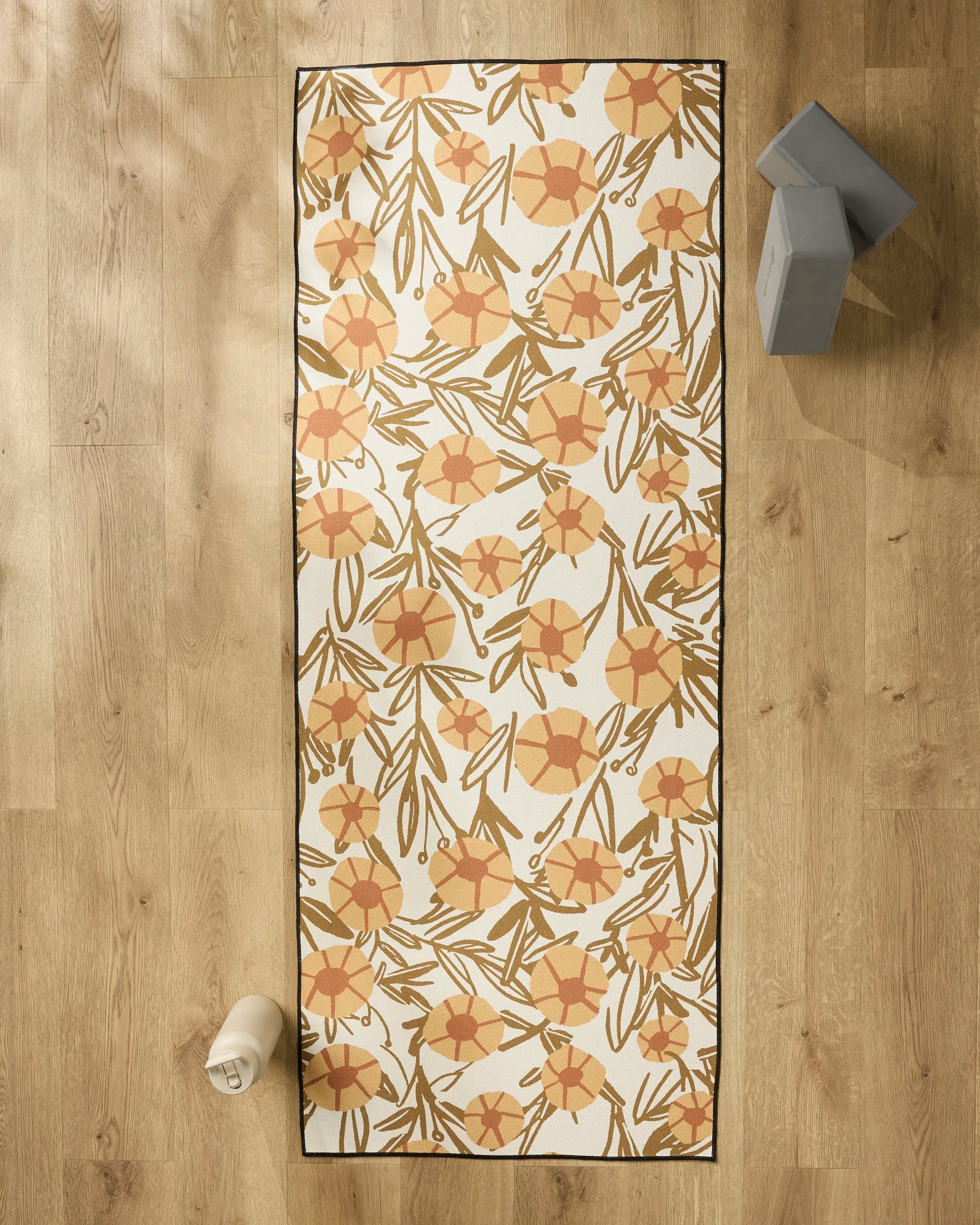 The "Sunflower Fields" yoga mat by Janna Sue Design with a floral orange and yellow pattern rests on a wooden floor, paired with a non-slip yoga towel, water bottle (bottom left), and two gray blocks (top right).