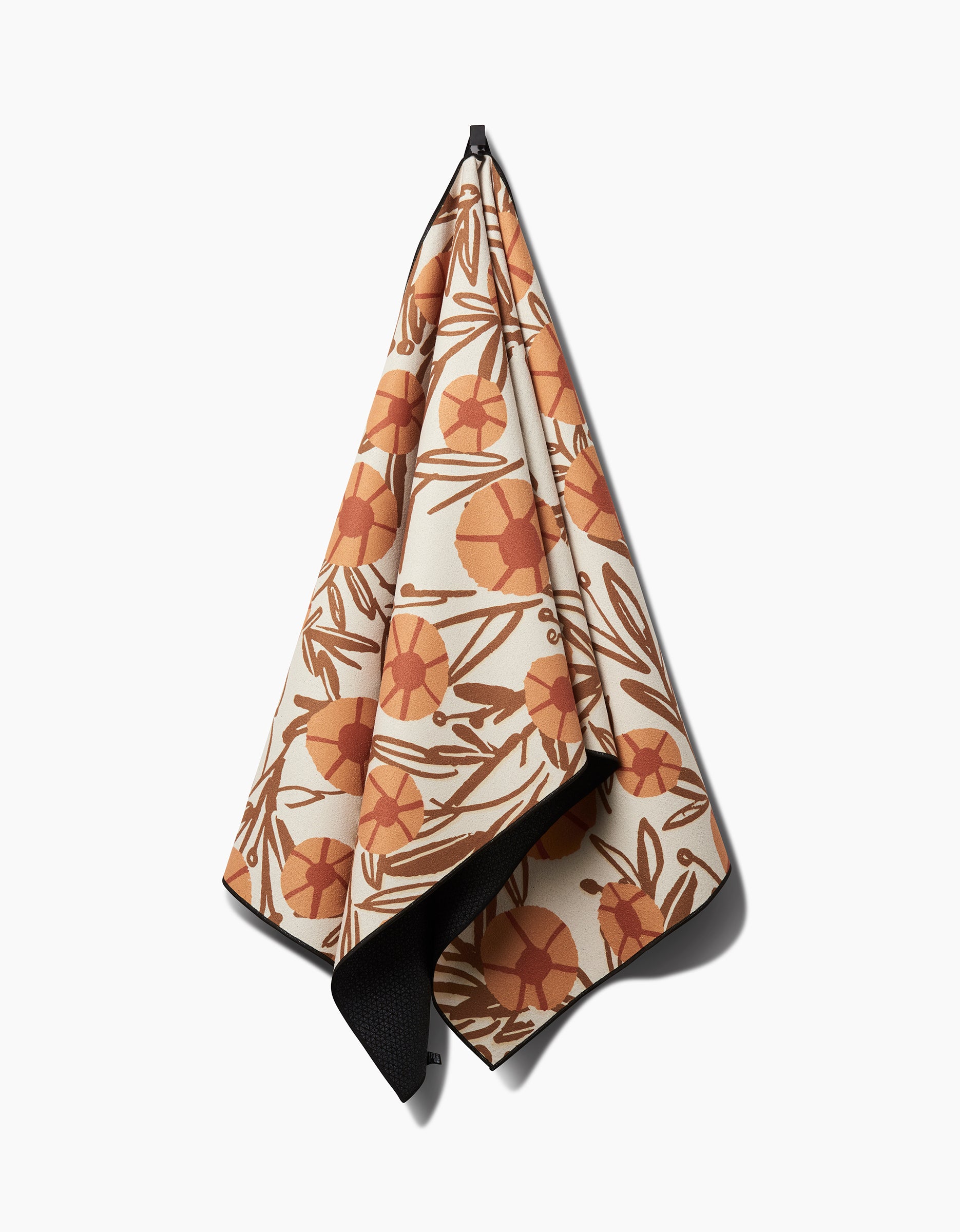 The Janna Sue Design Sunflower Fields yoga towel, featuring an orange and brown floral print, hangs on a hook. A corner reveals its black silicone-grip underside for stability against a white background.