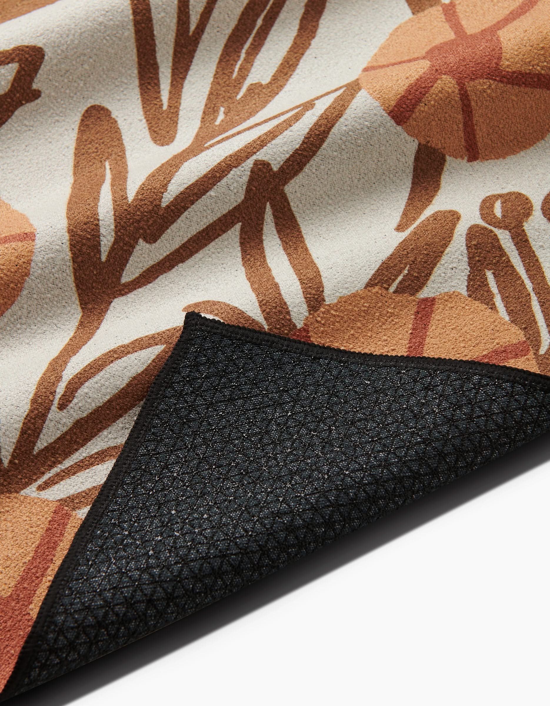A close-up of Sunflower Fields by Janna Sue Design shows its brown and orange floral pattern on cream, with the textured black silicone-grip underside slightly folded at the corner.