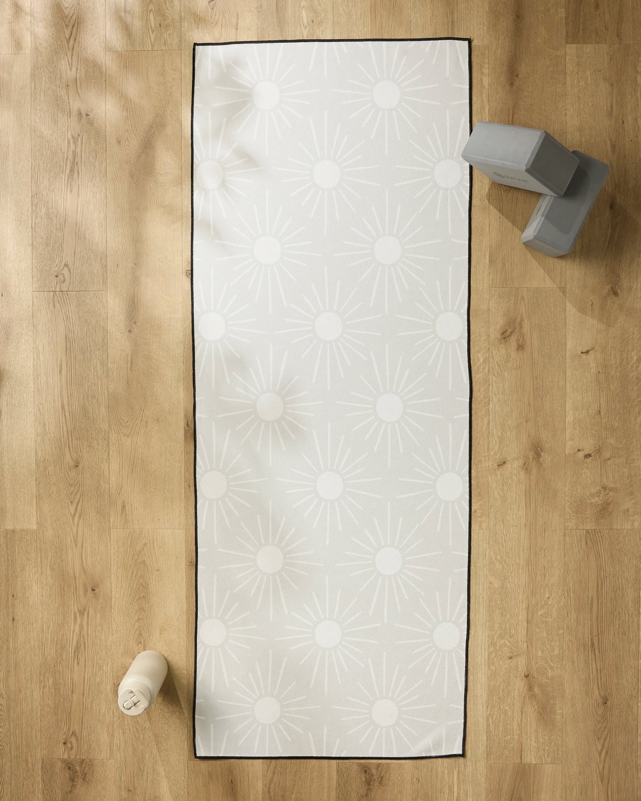 The Hufton Studio Sunny Geo white yoga mat with a sunburst pattern rests on a wooden floor, accompanied by a recycled yoga towel, water bottle at the lower left, and two stacked gray yoga blocks at the upper right.