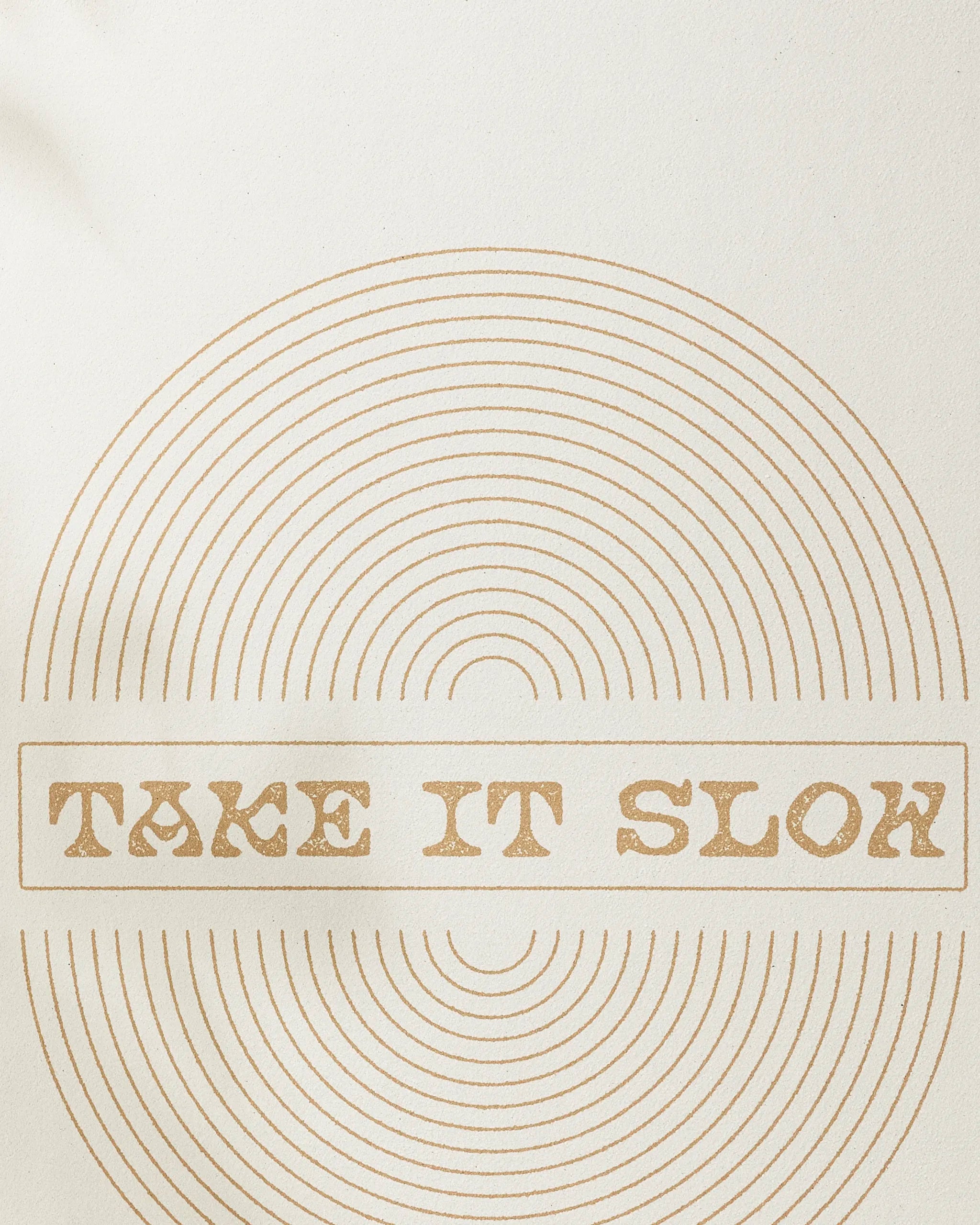 Beige background with concentric circles and “TAKE IT SLOW” in stylized brown text, centered in a rectangle—design by Graphics and Grain, inspired by the calming focus of their non-slip yoga towel.