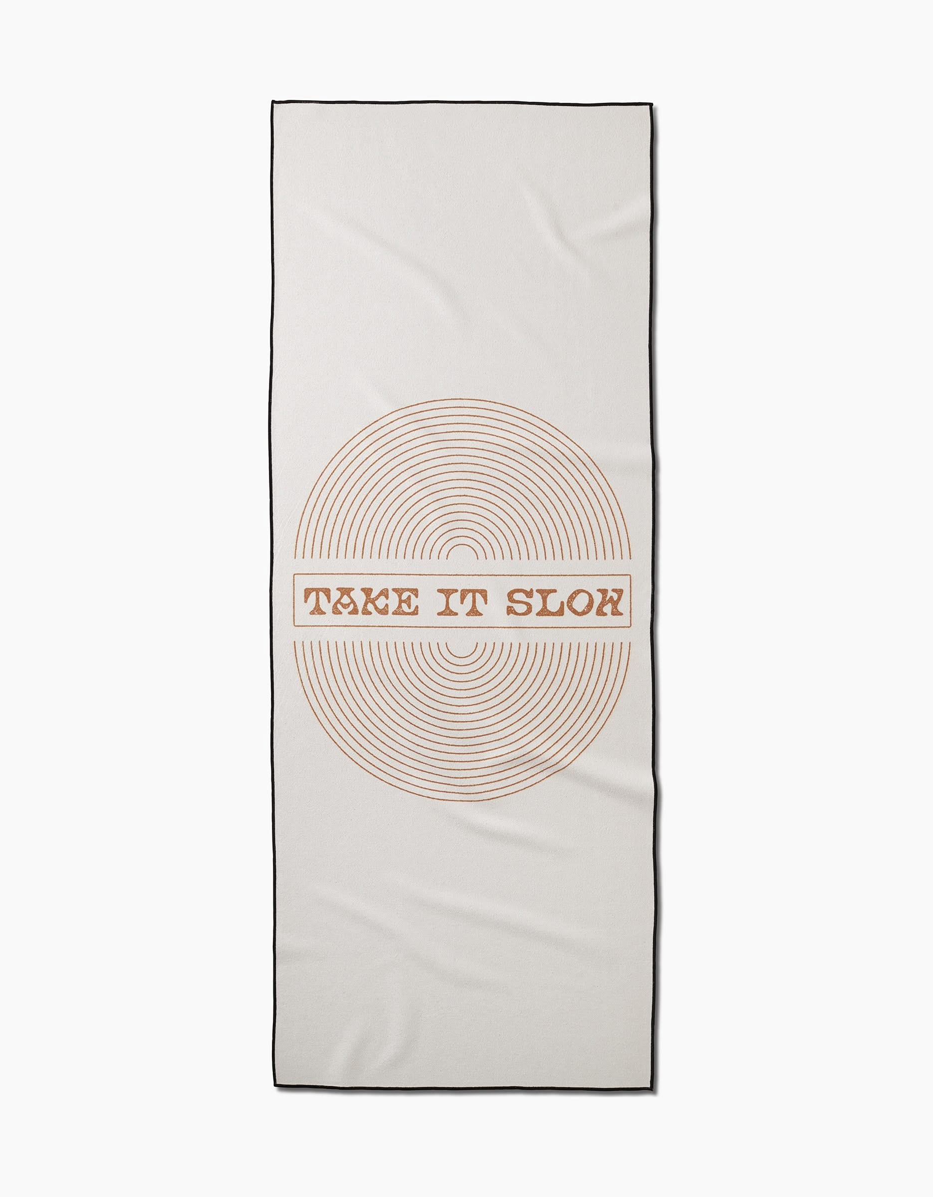 The Take It Slow yoga towel by Graphics and Grain is rectangular, white with black trim, and features a circular pattern of thin orange lines with bold retro text. Made from recycled materials for eco-friendly practice.