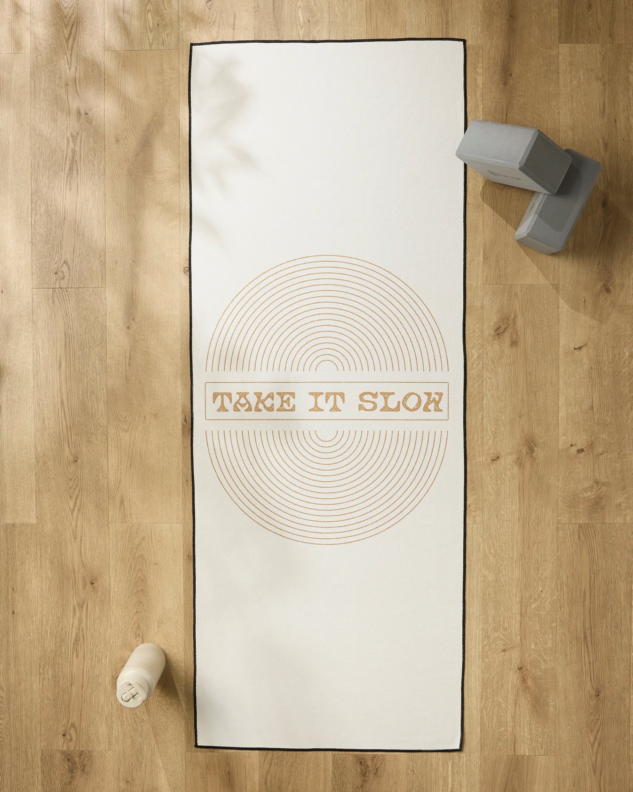 The "Take It Slow" yoga mat by Graphics and Grain features white concentric circles with the phrase TAKE IT SLOW in the center, set on a wooden floor alongside a non-slip towel, water bottle, and two gray yoga blocks.