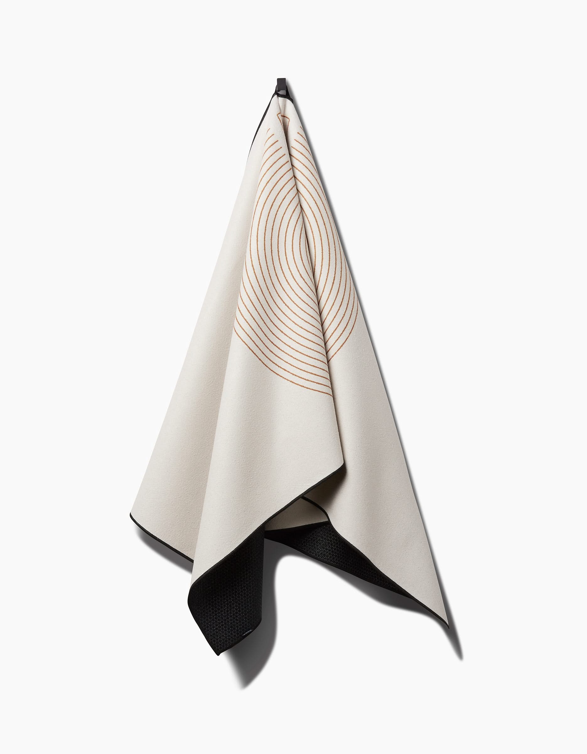 The Take It Slow yoga towel by Graphics and Grain hangs from a hook against a white background, showcasing cream fabric with orange concentric circles and a contrasting dark underside for a simple, modern look.