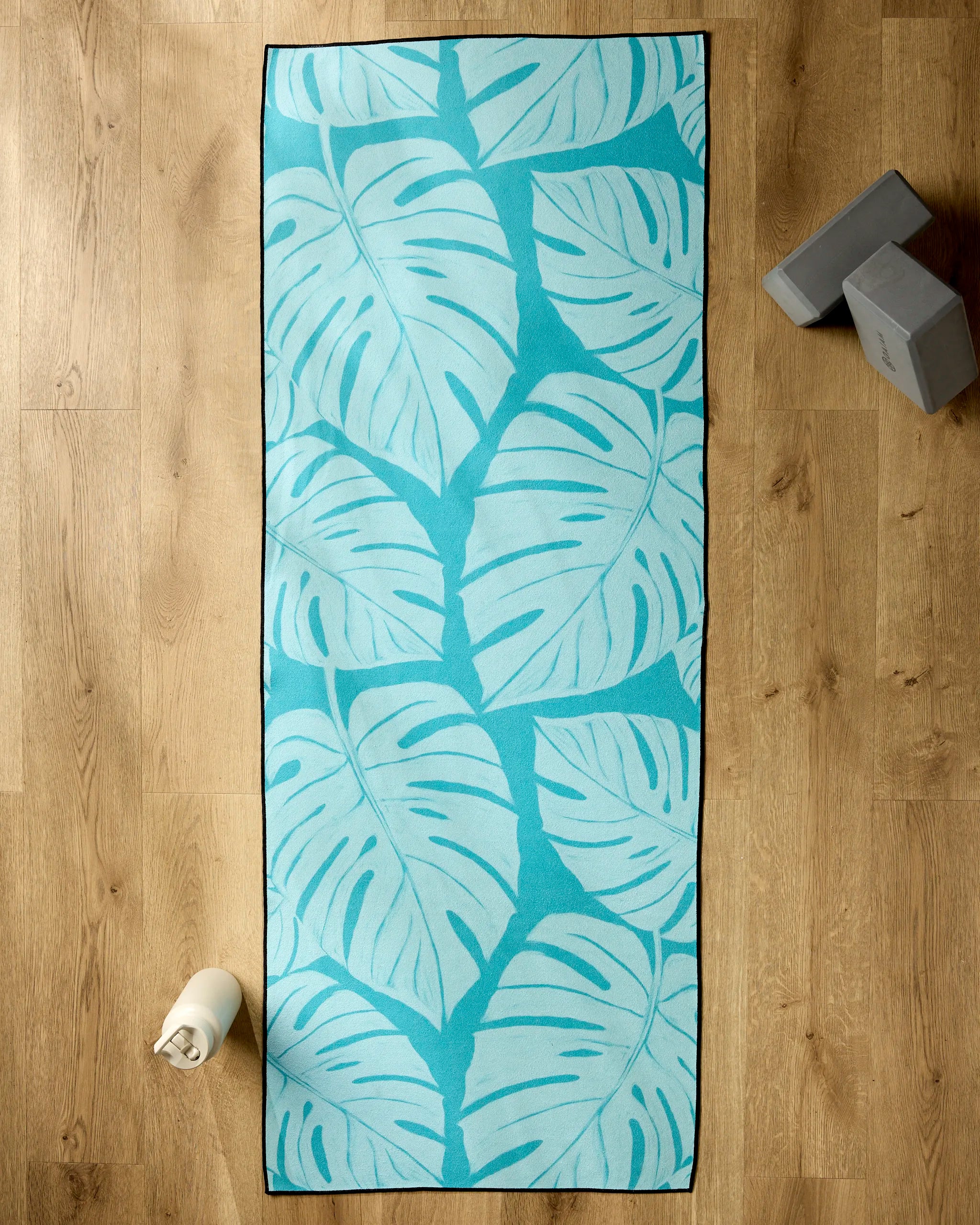 The GEOMETRY Tropical yoga mat, featuring a large turquoise tropical leaf design, rests on a wooden floor with a water bottle, two gray yoga blocks, and a non-slip yoga towel arranged nearby.