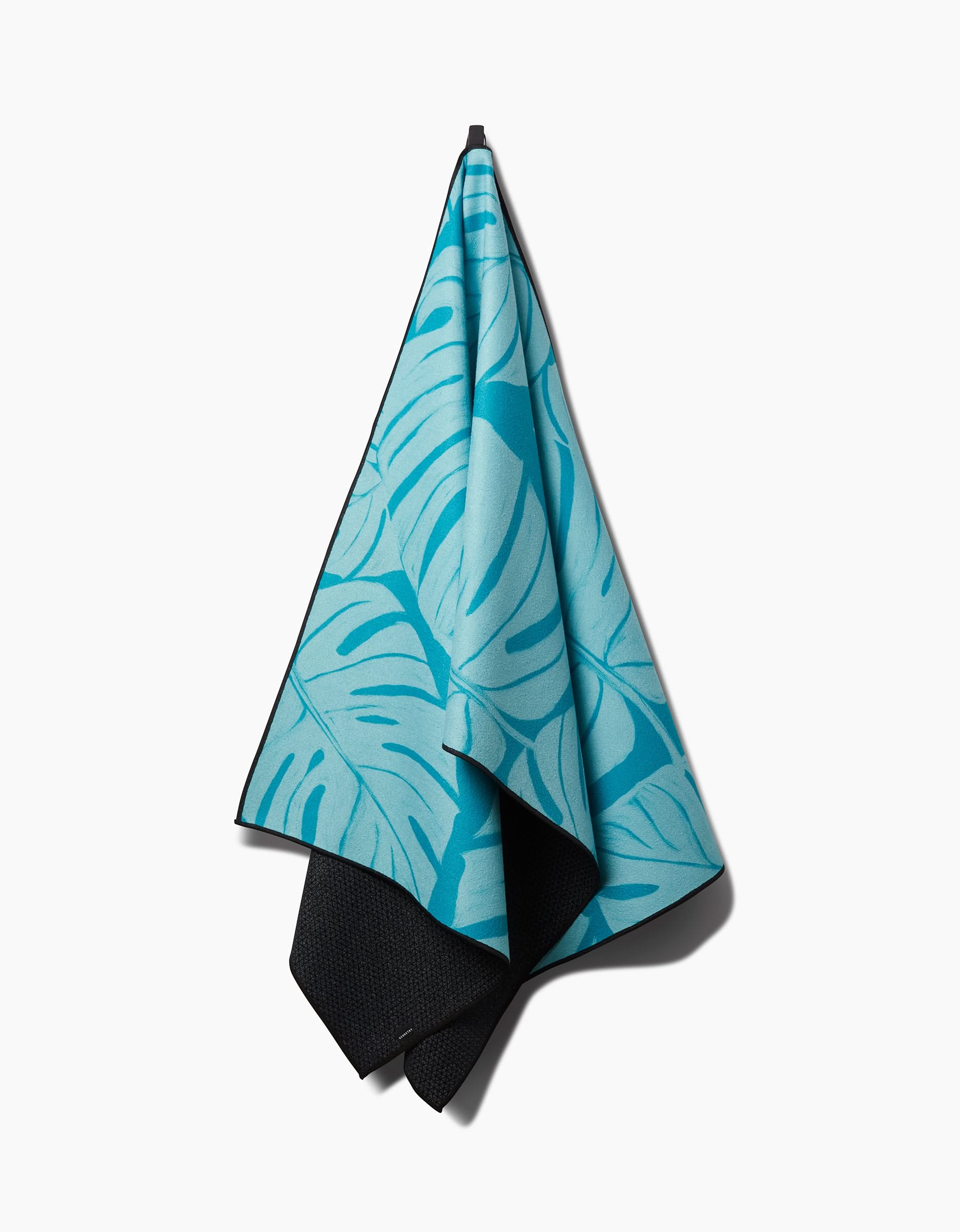 The GEOMETRY Tropical yoga towel features a blue tropical leaf pattern and hangs on a hook against a white background. Its black non-slip underside adds both style and stability to your practice.