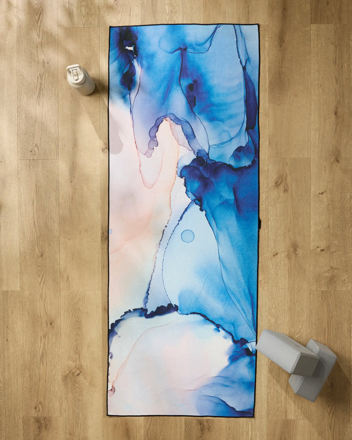 The GEOMETRY Water And Ink non-slip yoga towel, featuring a blue and light pink abstract watercolor design, is displayed on a wooden floor with a water bottle and two yoga blocks.