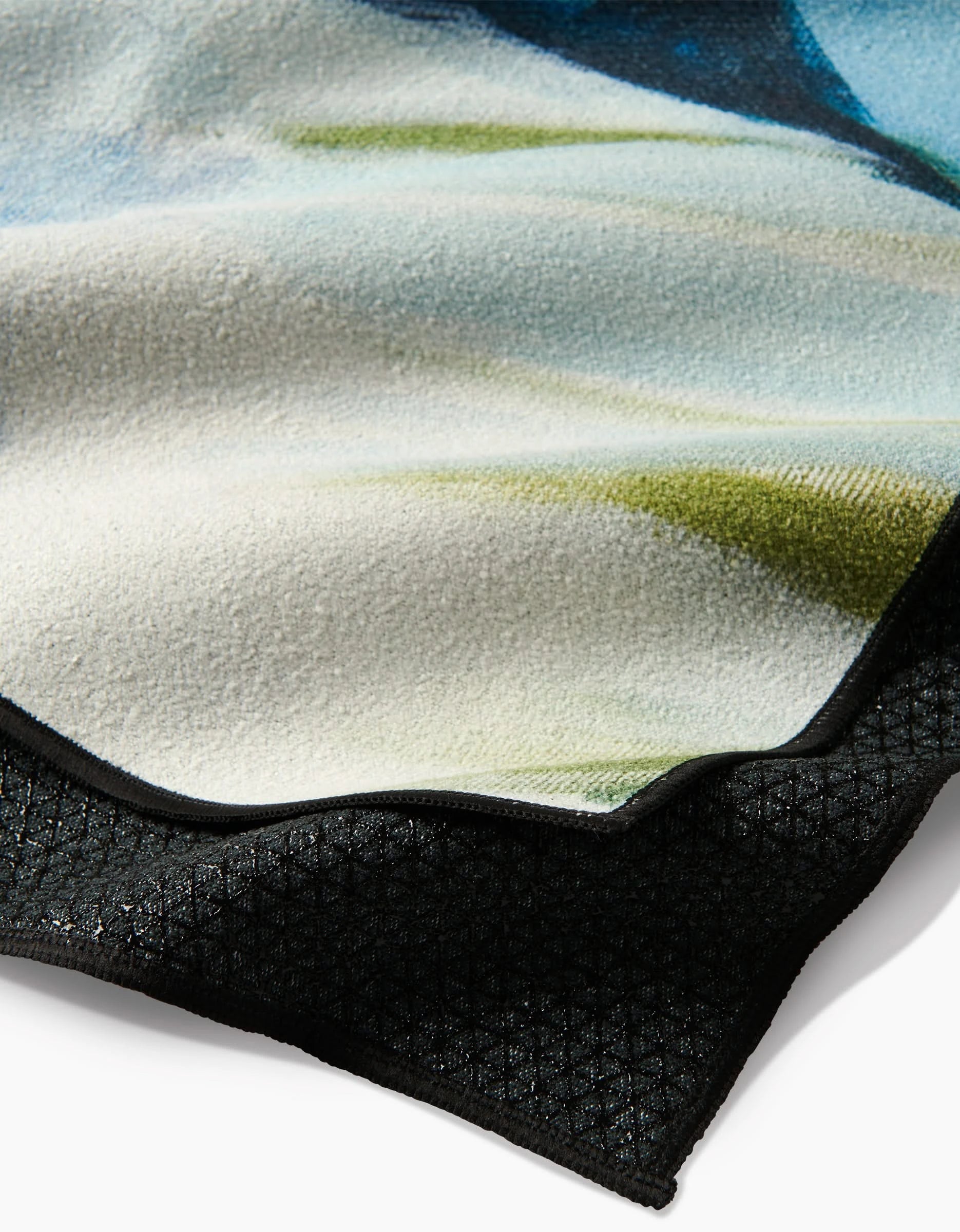 A detailed close-up of the Where He Laid To Go yoga towel by Kelly Peters, crafted from recycled materials with a white, green, blue, and black printed pattern, folded edge, and fine stitching.
