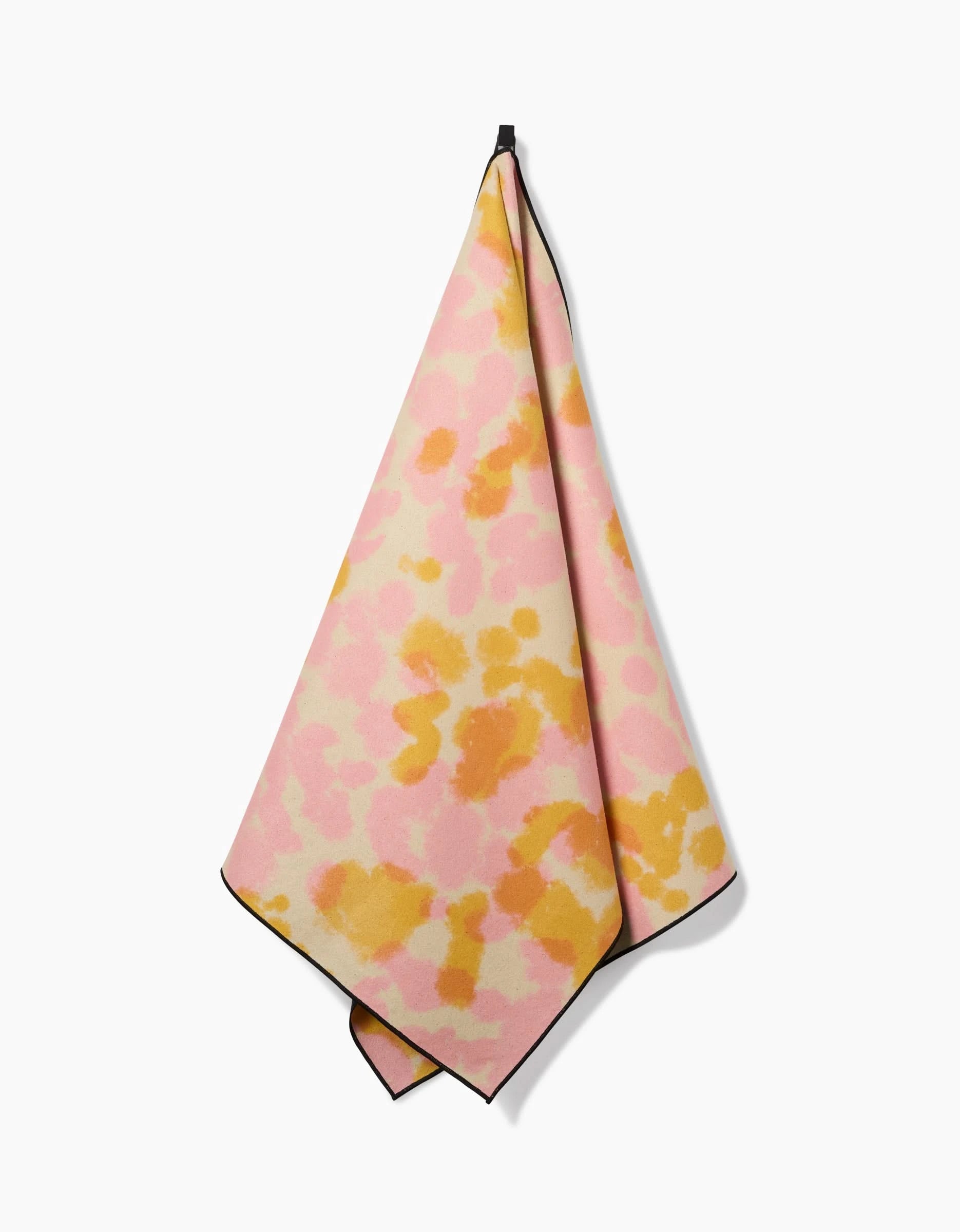 The GEOMETRY Whimsy yoga towel, featuring a pink, yellow, and orange abstract watercolor design and made from recycled materials, hangs by a black loop against a white background.