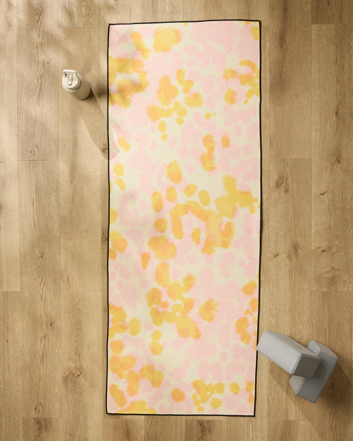 The GEOMETRY Whimsy yoga mat, featuring a pastel pink and yellow abstract pattern, is laid on a wooden floor with a non-slip towel. Alongside it are a white water bottle and gray yoga blocks.