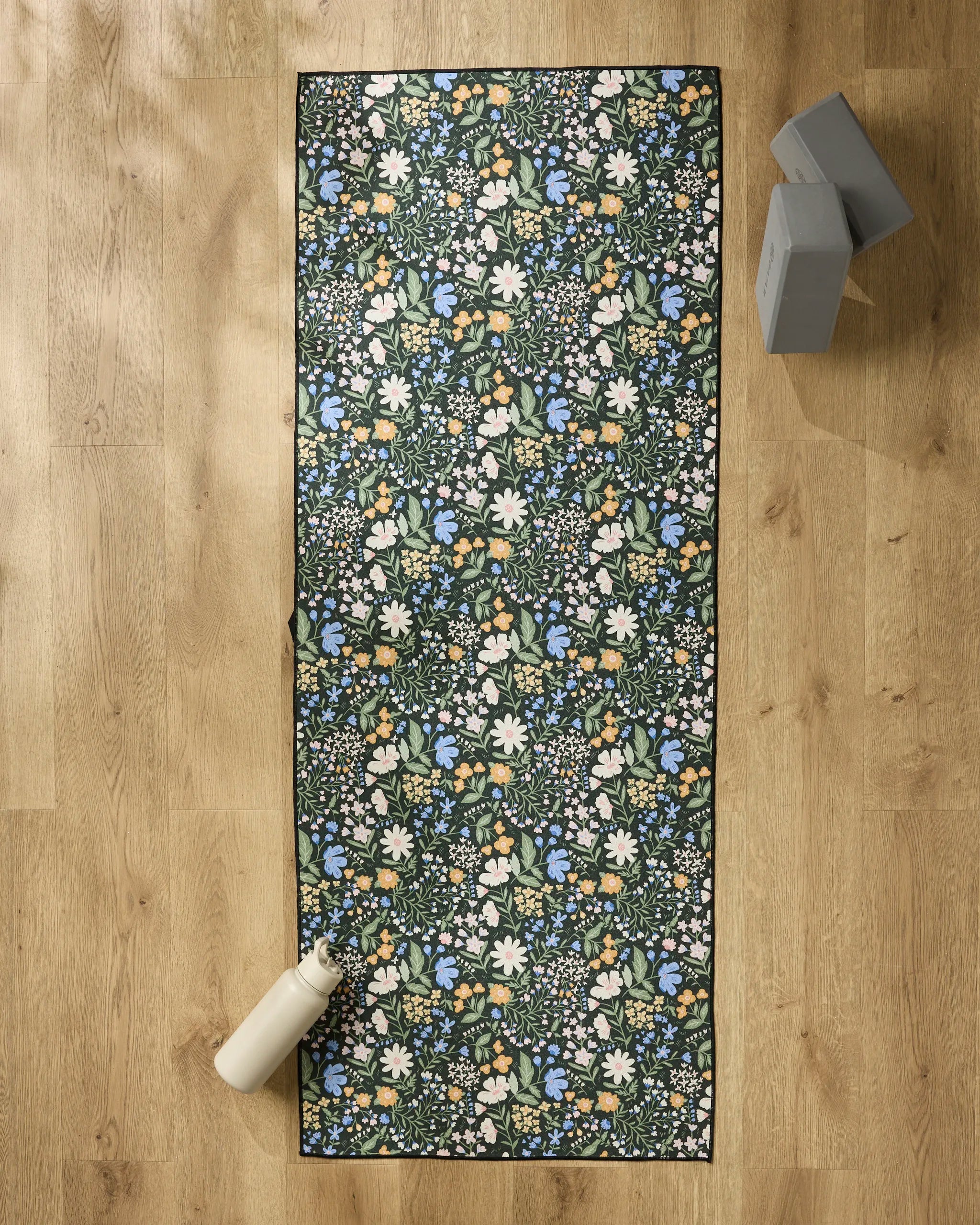 The Danelys Sidron Wildflower Vine Blush yoga mat, featuring white, blue, and yellow flowers, rests on a light wooden floor with a non slip towel, a white water bottle at one end, and two gray yoga blocks nearby.