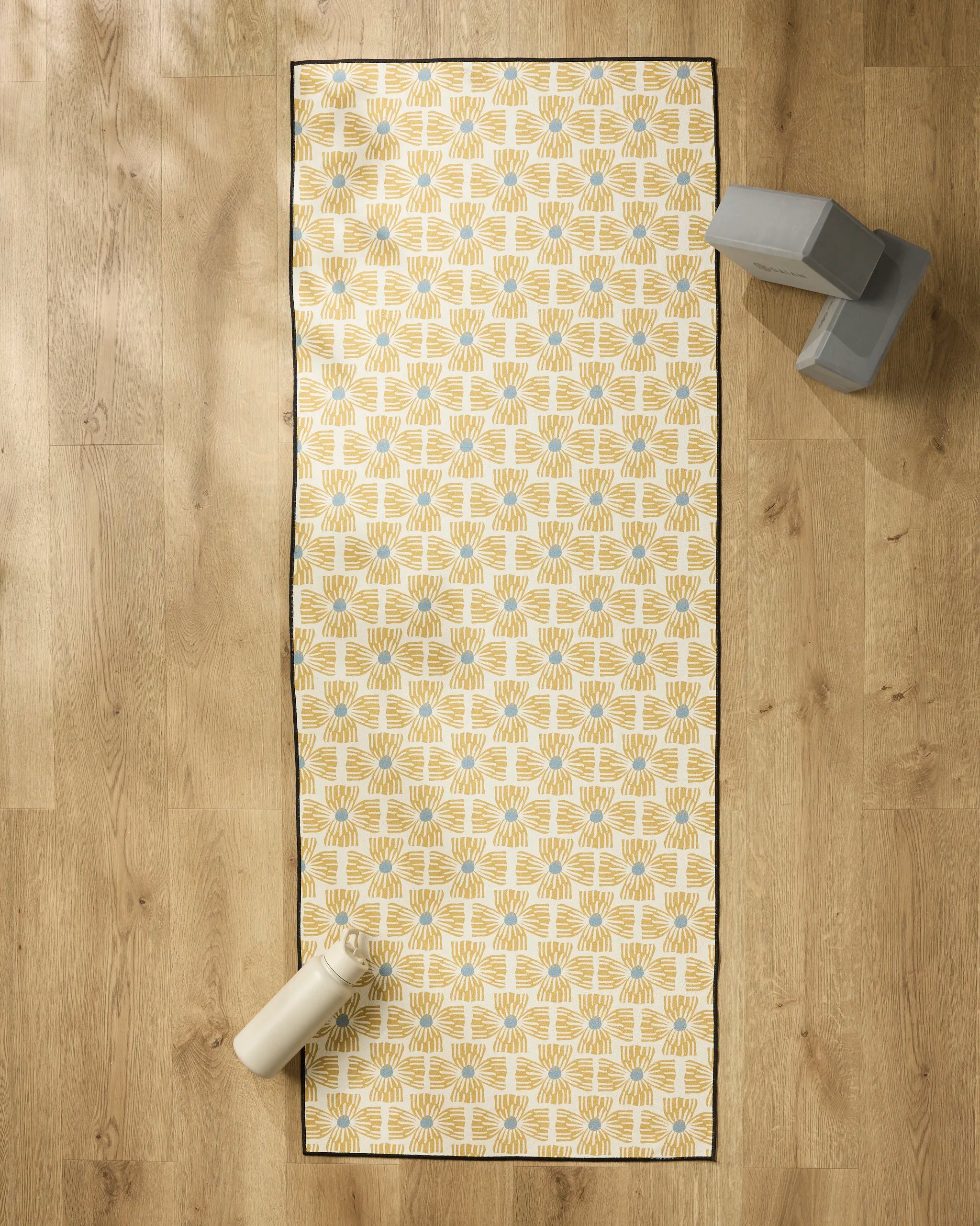 The Ash Cascade Wildflower non-slip yoga towel with a yellow and white pattern is on a wooden floor, a white water bottle in the bottom left, and two gray yoga blocks in the top right corner.