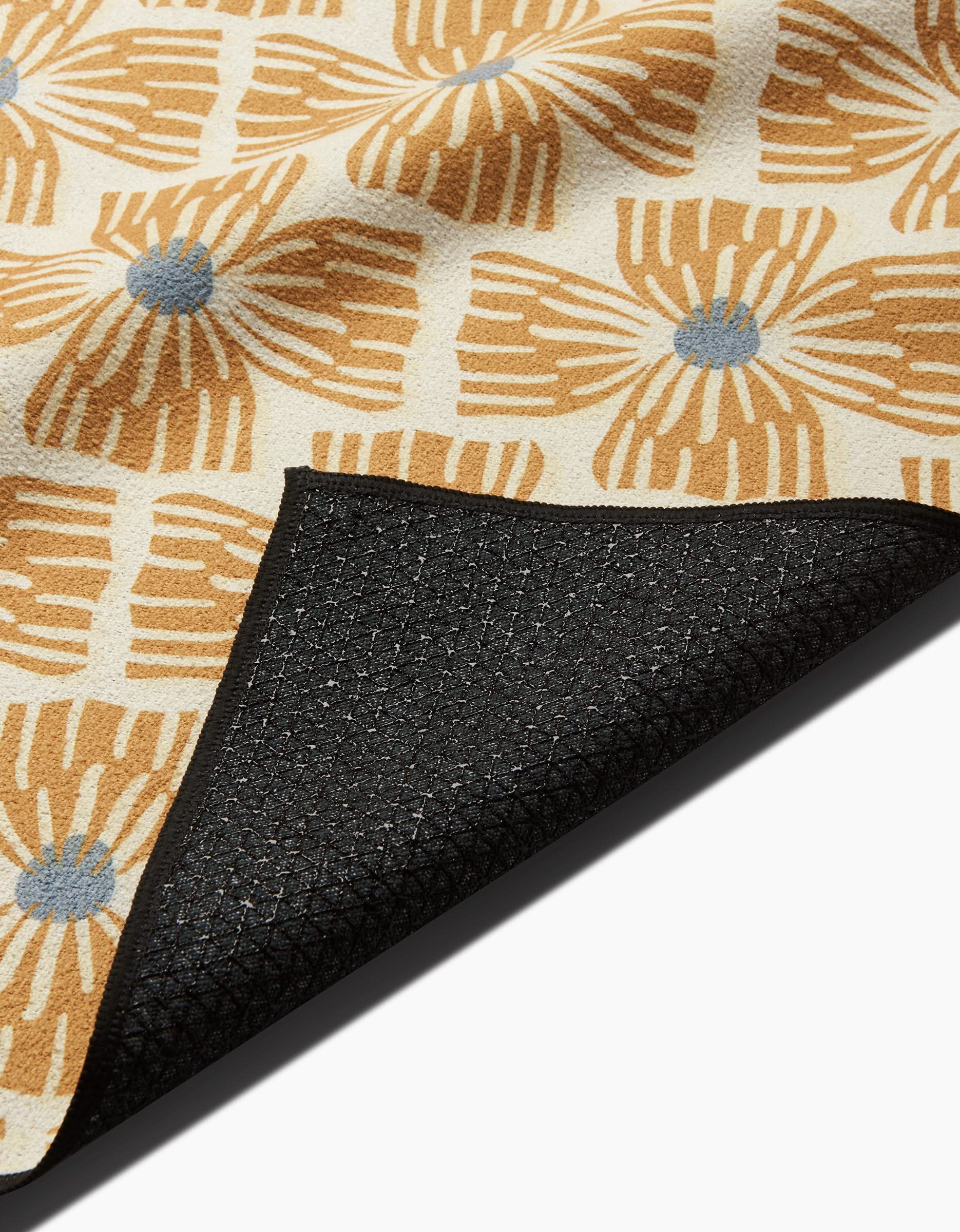 A close-up of the Ash Cascade Wildflower yoga towel shows its tan and white floral pattern with blue centers, and one corner folded over to display the black textured, slip-resistant backing.