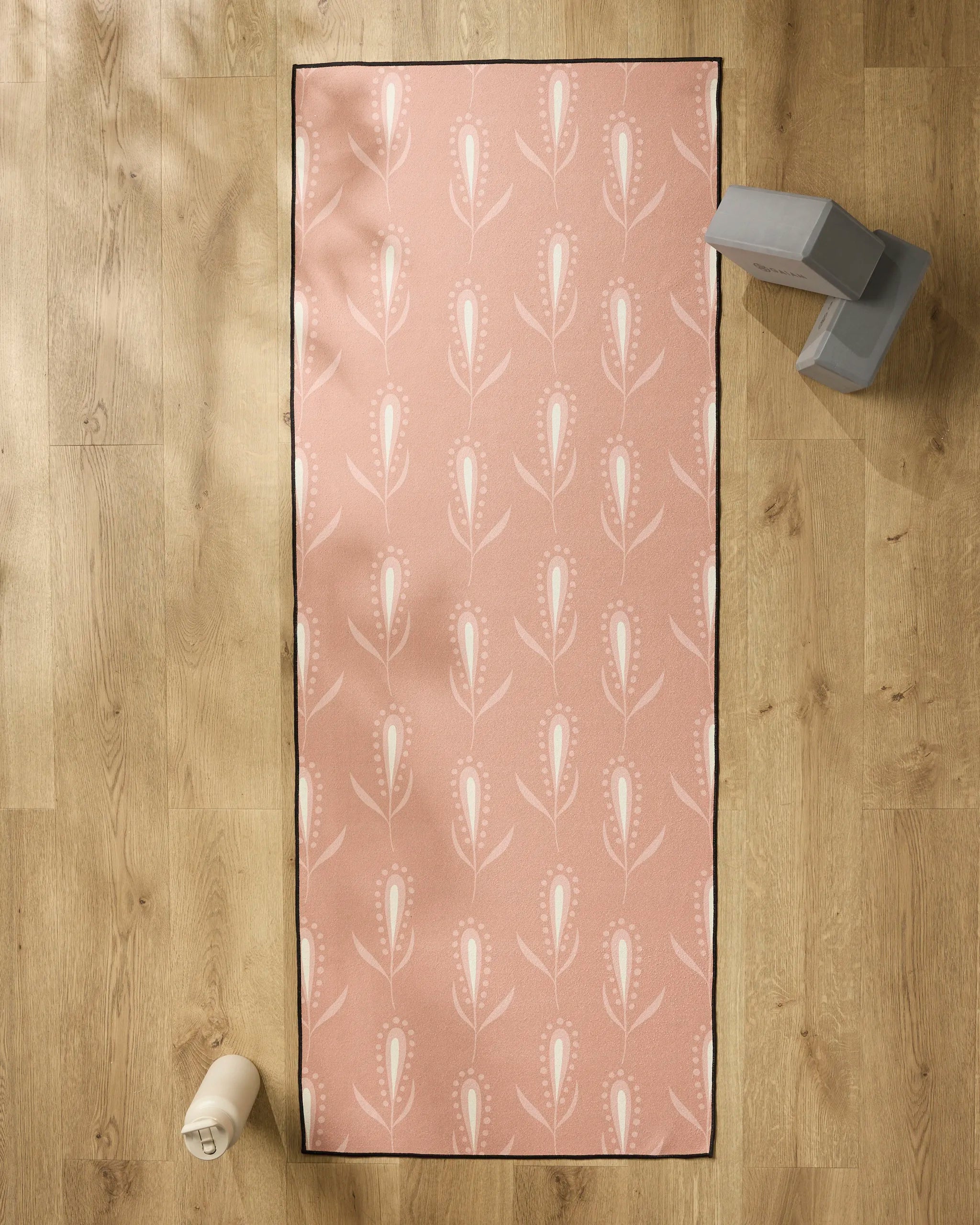 The Amita Nair Wilds pink yoga mat with a white floral pattern is laid out on a wooden floor, topped with a non-slip yoga towel. A water bottle sits to the left, and gray exercise blocks rest at the top right of the mat.