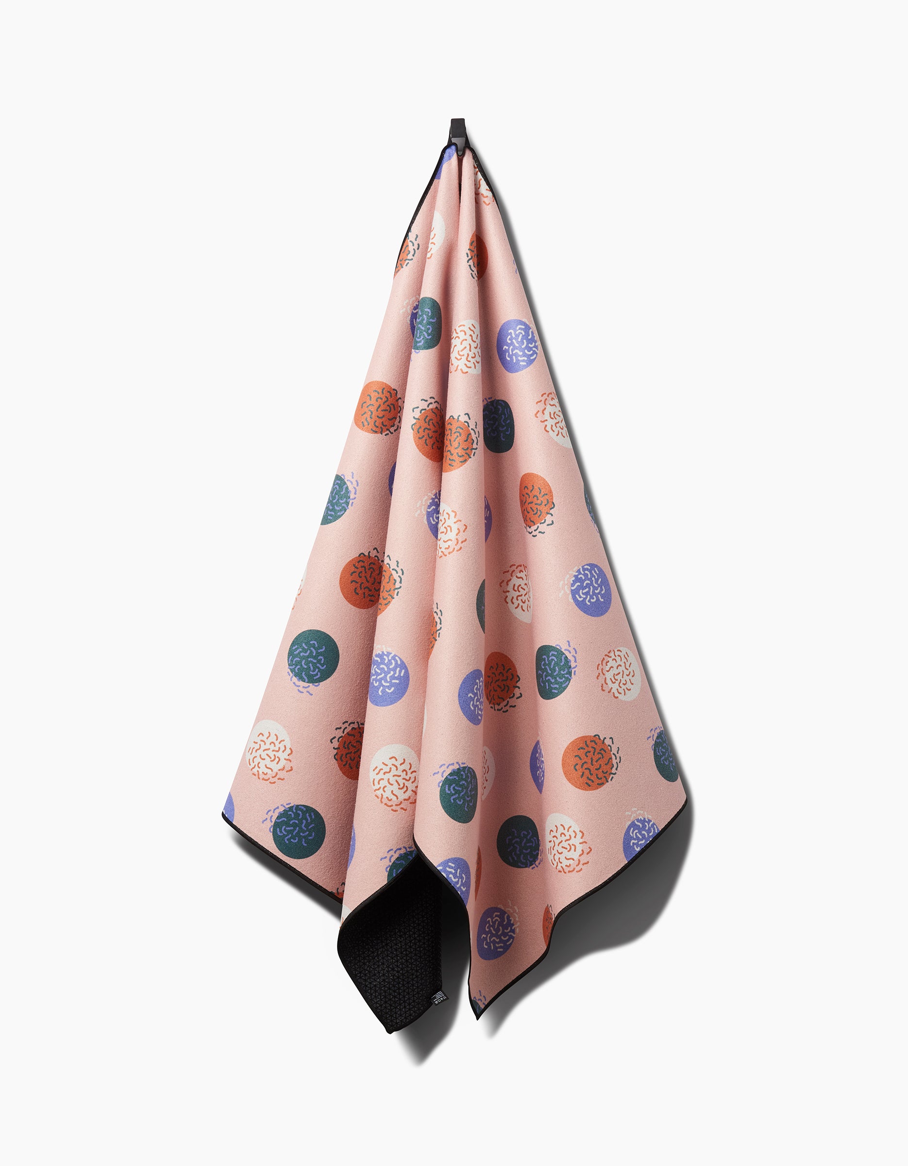 The Claire Iglesias Wonky Dots yoga towel, peach-colored with multicolored textured circles, is hanging from a hook against a white background.