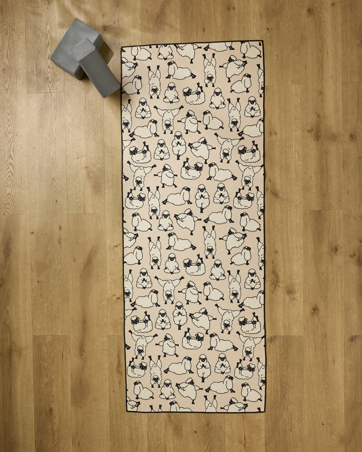 The Geometry Yoga Sheep mat, featuring a black and white cartoon dog design, lies on a wooden floor with a gray yoga block at the top left and a silicone grip towel nearby to support your practice.