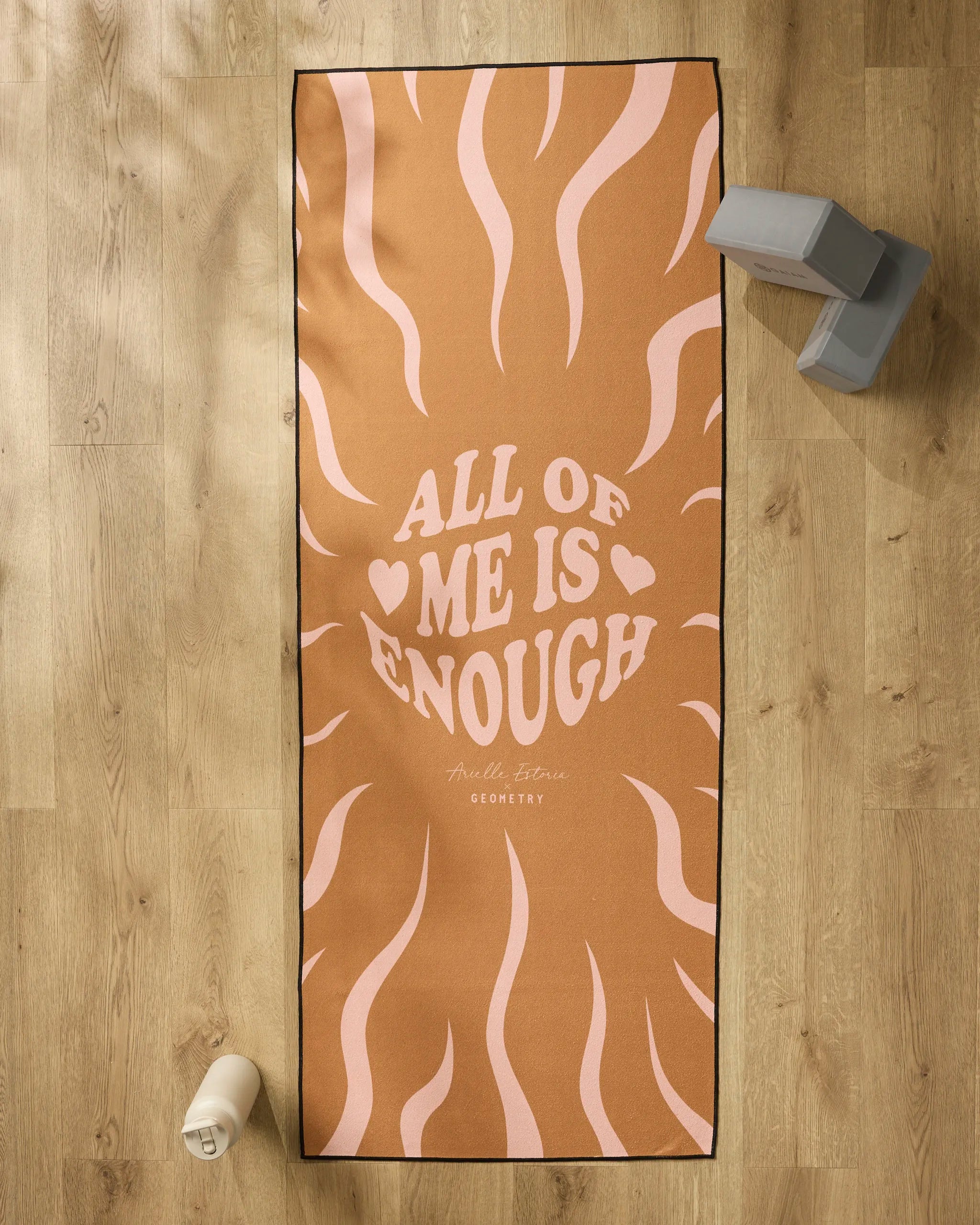 The “You are ENOUGH” yoga mat by Arielle Estoria features wavy pink lines and bold lettering, shown on a wooden floor with a silicone-grip yoga towel, water bottle, and two yoga blocks nearby.