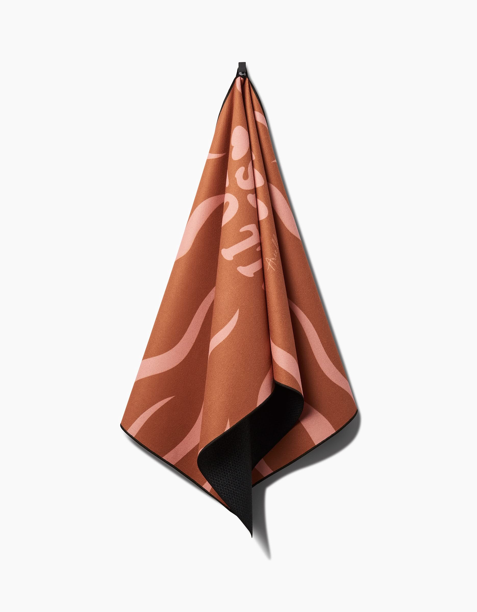 The You are ENOUGH brown yoga towel by Arielle Estoria, featuring pink abstract patterns and lettering on recycled material, hangs from a hook against a plain white background.