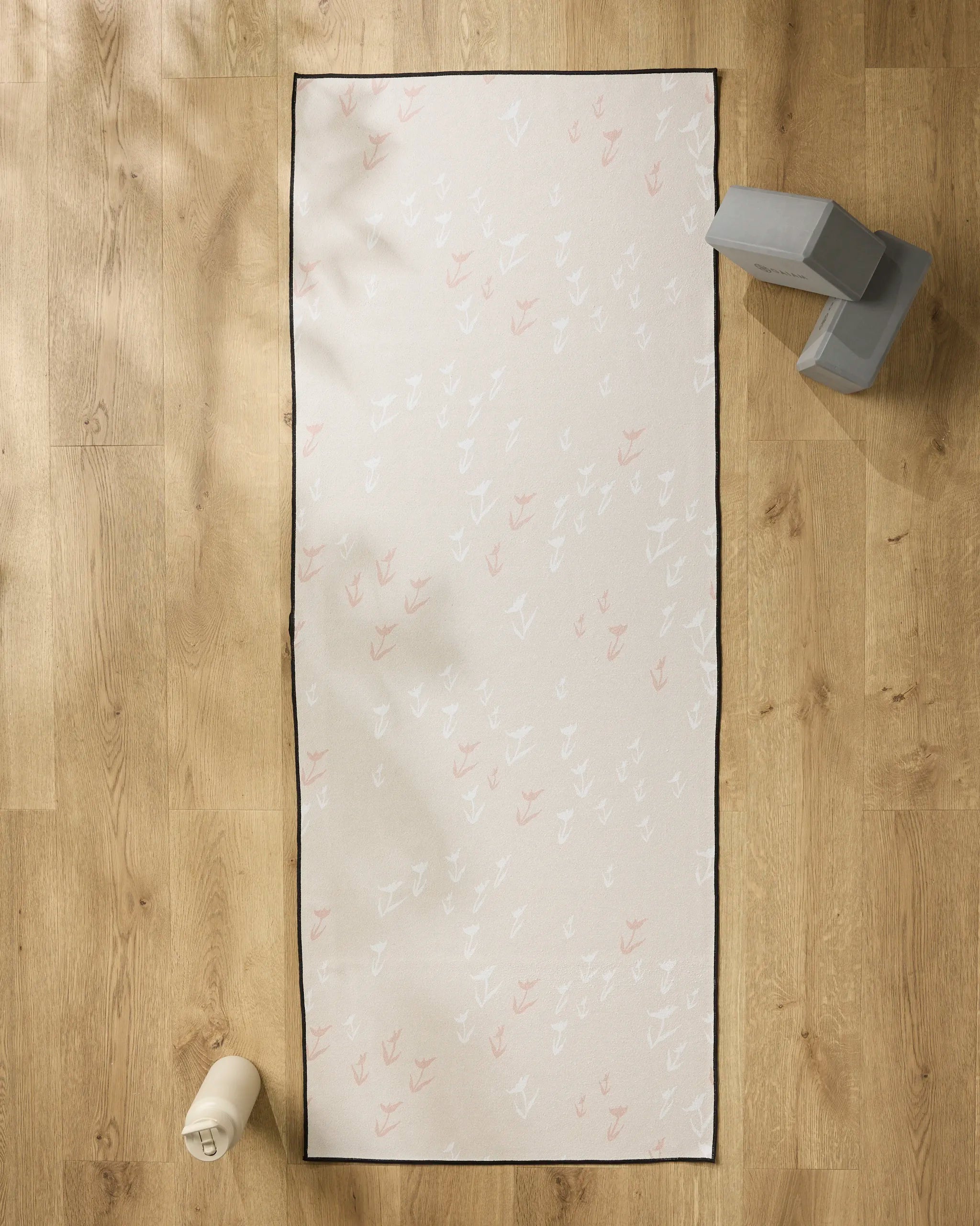 The You Grow Girl yoga mat by Aubrey Fairchild, featuring a white and pink feather pattern, lies on a light wood floor with a slip-resistant towel. Nearby are gray yoga blocks and a white water bottle.
