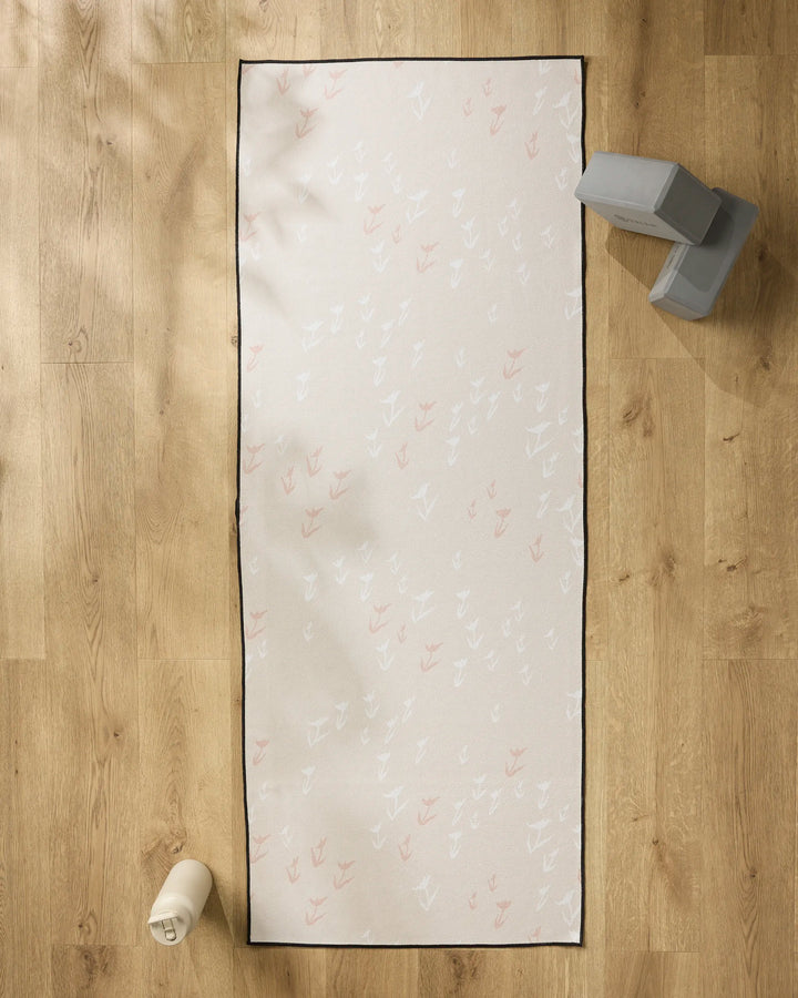 The You Grow Girl yoga mat by Aubrey Fairchild, featuring a white and pink feather pattern, lies on a light wood floor with a slip-resistant towel. Nearby are gray yoga blocks and a white water bottle.