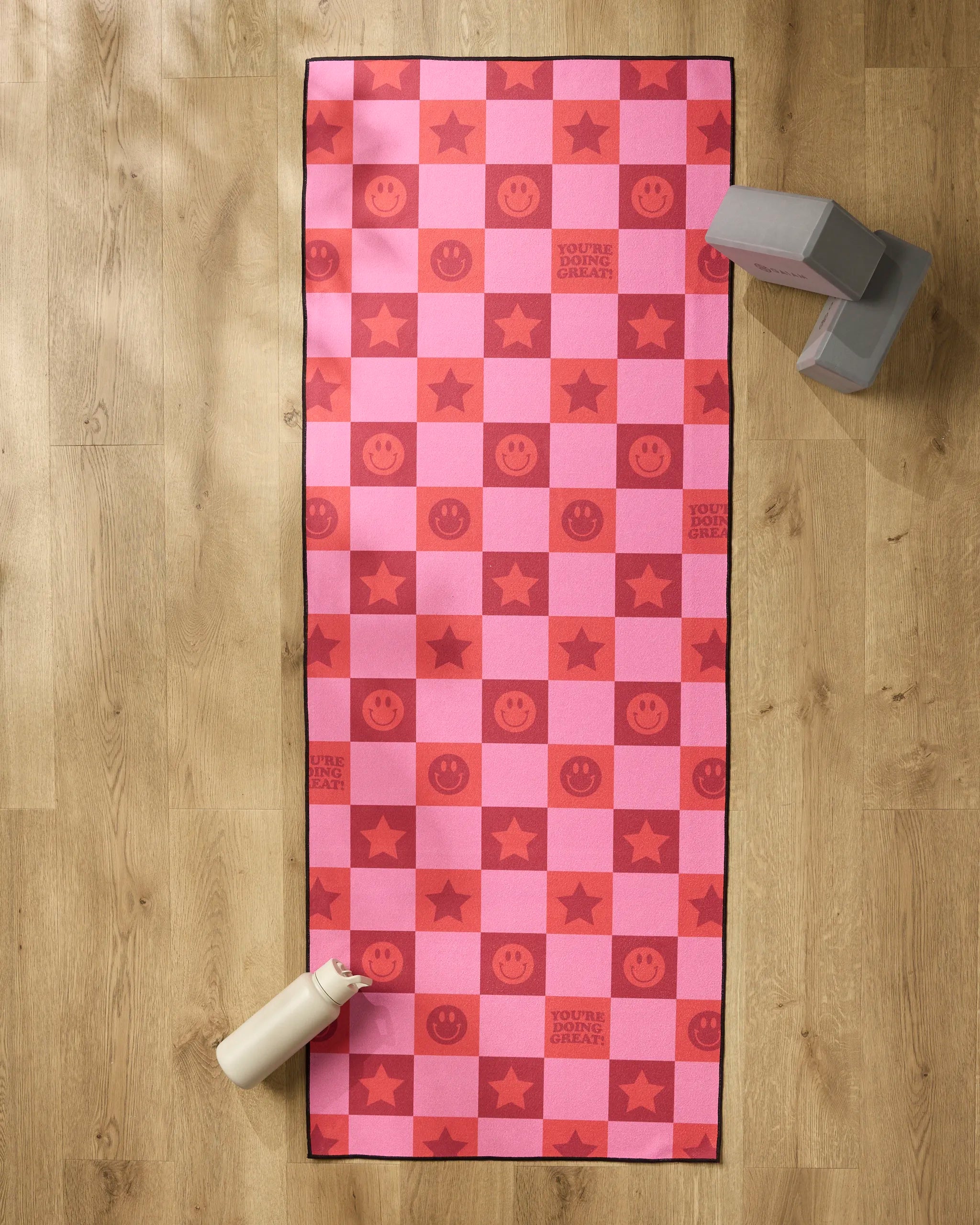The "You're Doing Great" yoga mat by Quotes by Christie features pink and red checkers, smiley faces, stars, and uplifting text. A white water bottle, recycled towel, and two gray blocks sit on a light wooden floor around the mat.