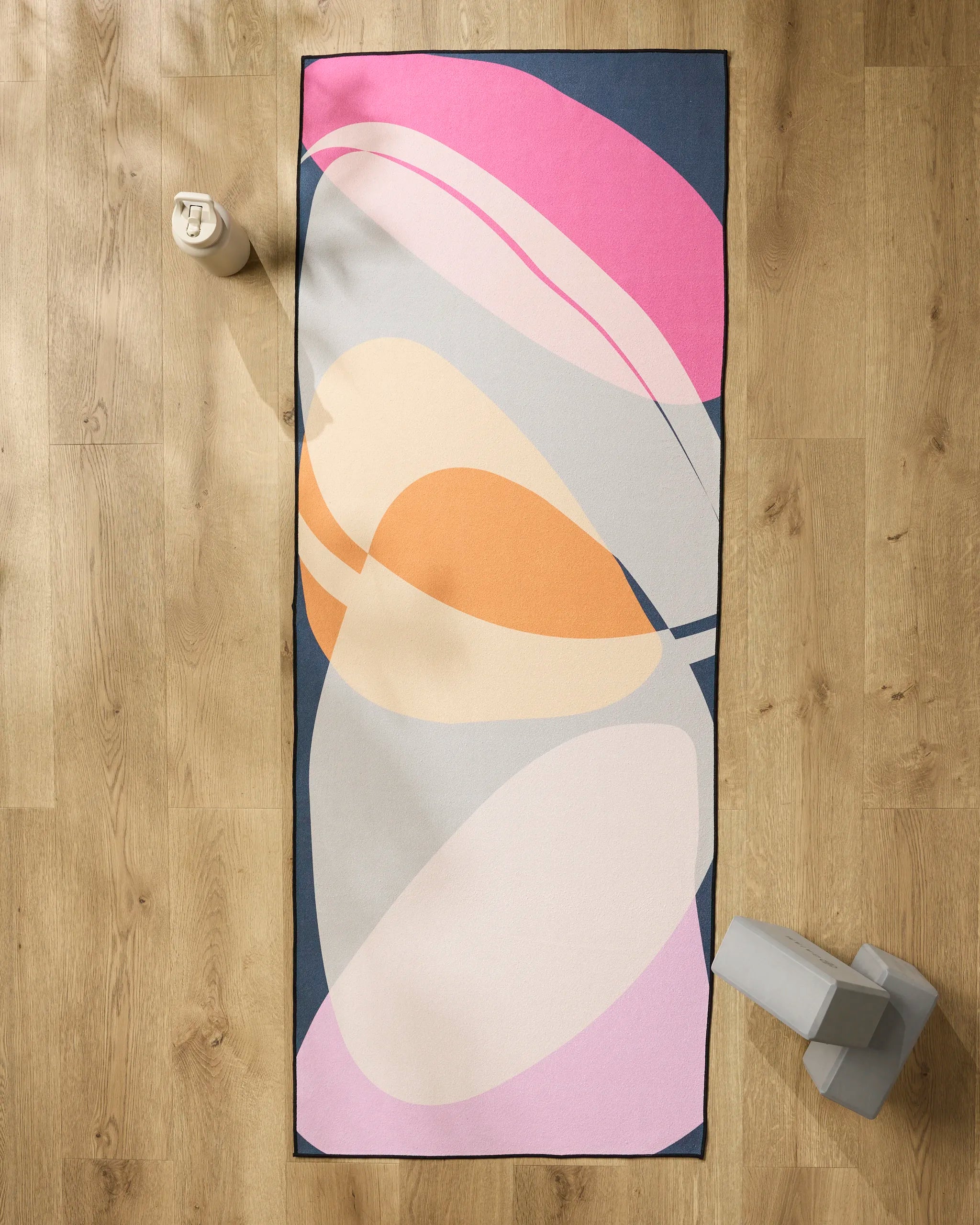 The GEOMETRY "Zen Summer" yoga mat, featuring abstract pink, orange, cream, and gray shapes, rests on a wooden floor. A silicone grip towel, water bottle (top left), and two gray yoga blocks (bottom right) complete the scene.