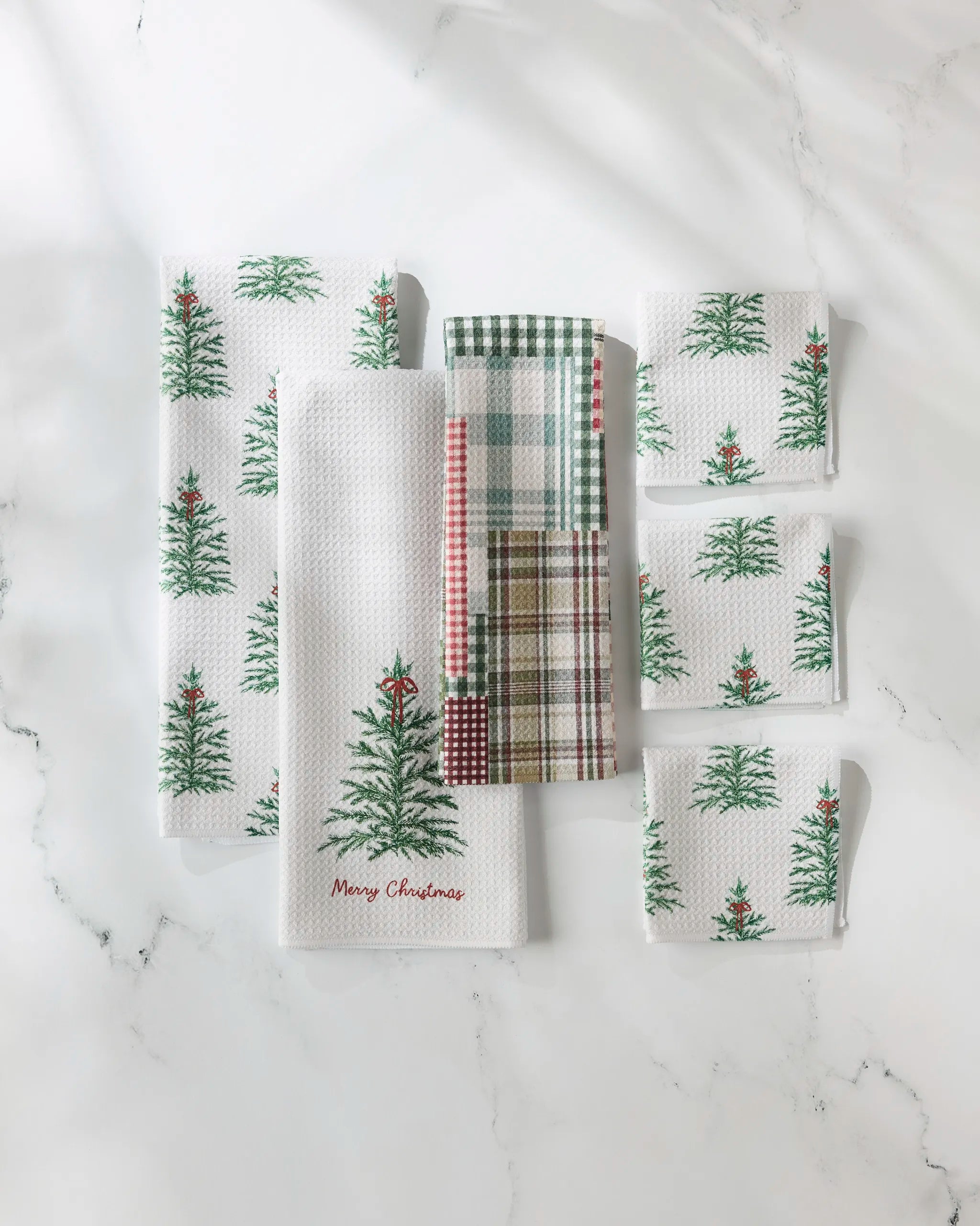Six GEOMETRY Redford kitchen towels—six white with green Christmas trees and red stars, two plaid in green, red, white, and beige—are arranged on a white marble surface. One towel features the words “Merry Christmas.”.
