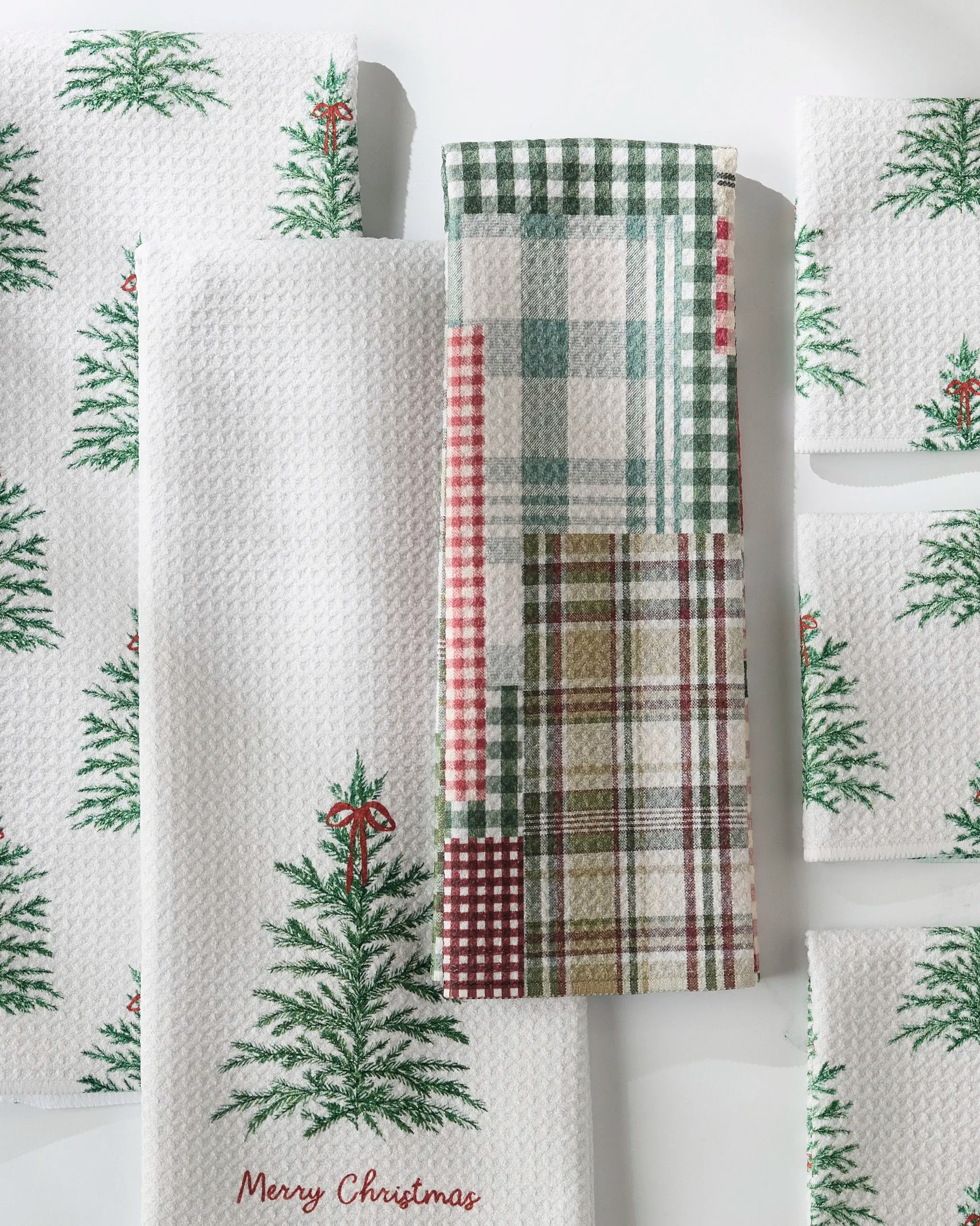 GEOMETRY Redford dish towels, featuring white fabric with green Christmas tree prints and a multicolored plaid design, are arranged neatly. One towel displays "Merry Christmas" in red at the bottom.
