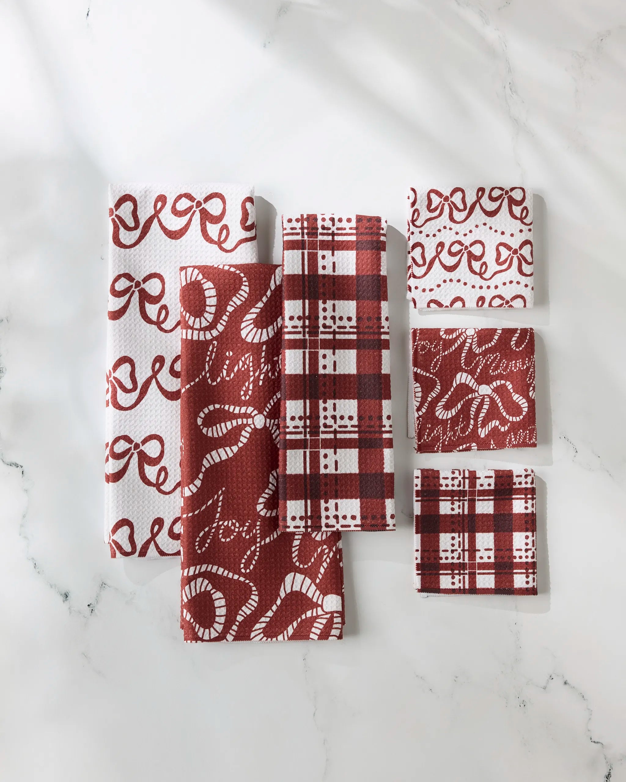 The GEOMETRY "Cheerful Bows" set includes seven red and white kitchen towels in assorted designs like plaid and cursive, all displayed on a white marble surface.
