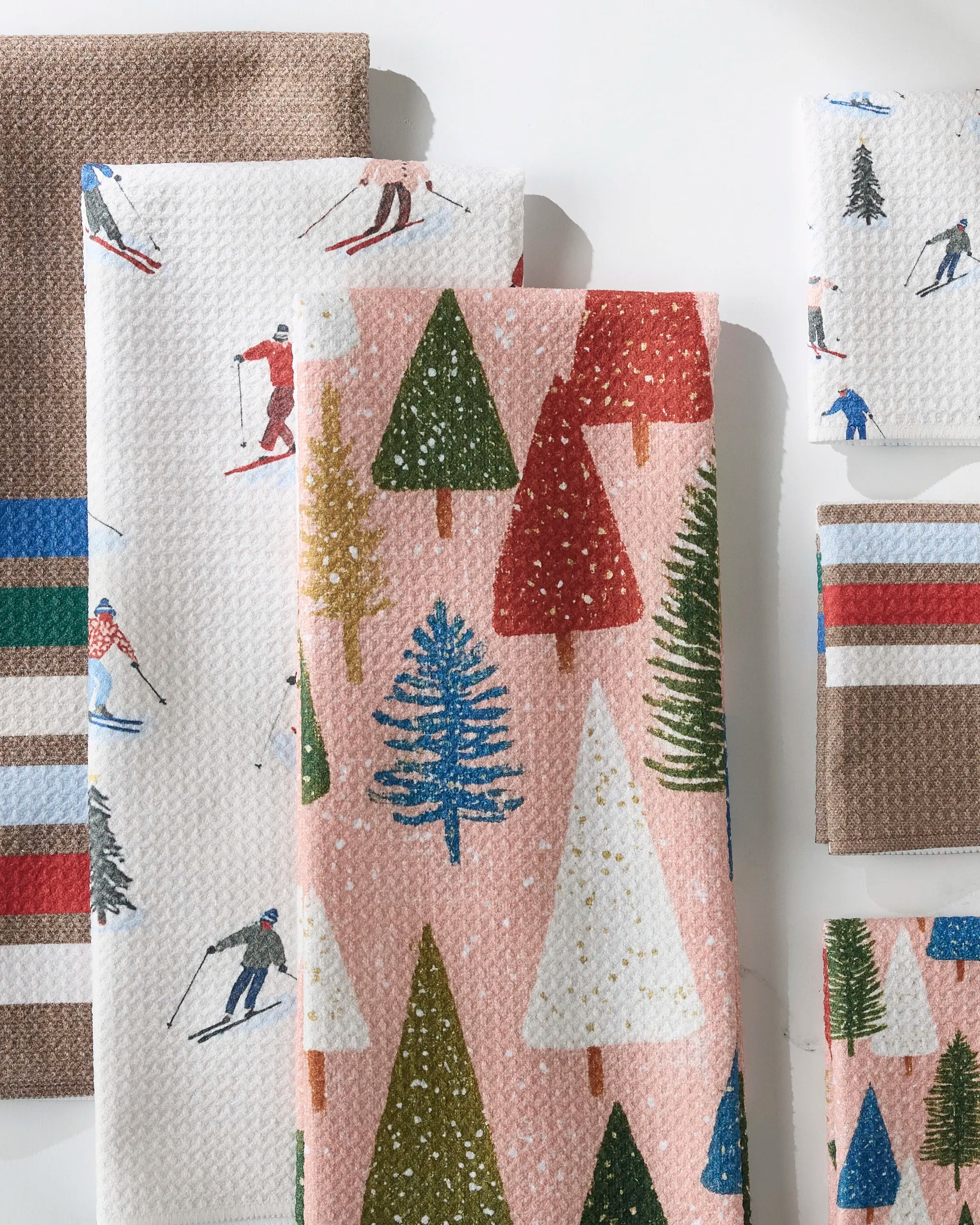 Several folded GEOMETRY Snowbound kitchen towels, featuring winter-themed designs like skiers, snowy trees, and vibrant stripes in pink, brown, green, red, and blue, are arranged on a white surface.
