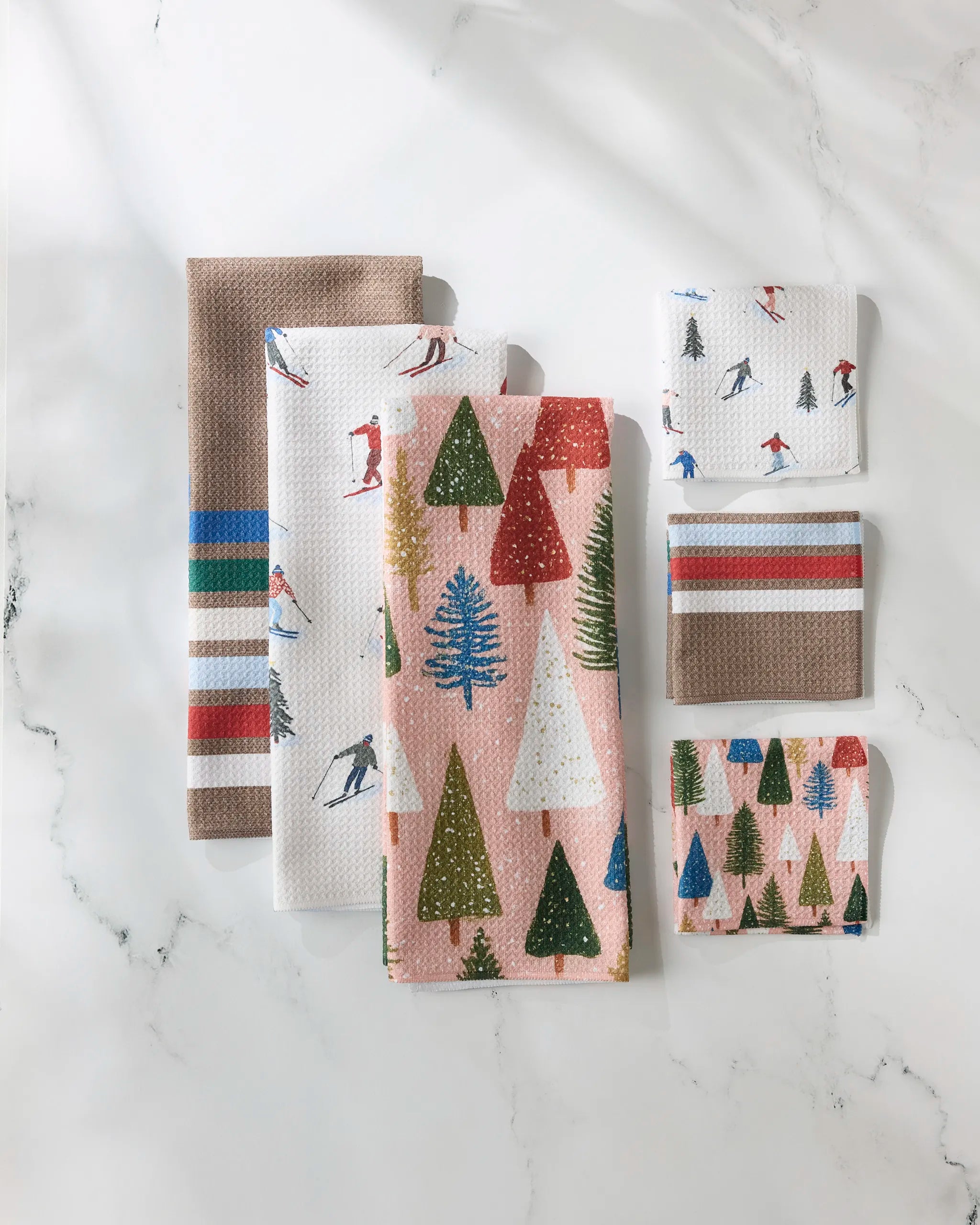 Snowbound by GEOMETRY: a festive set of kitchen towels and cloths featuring tree, skier, colorful stripe, and solid brown designs in large and small sizes, beautifully arranged on a white marble surface.