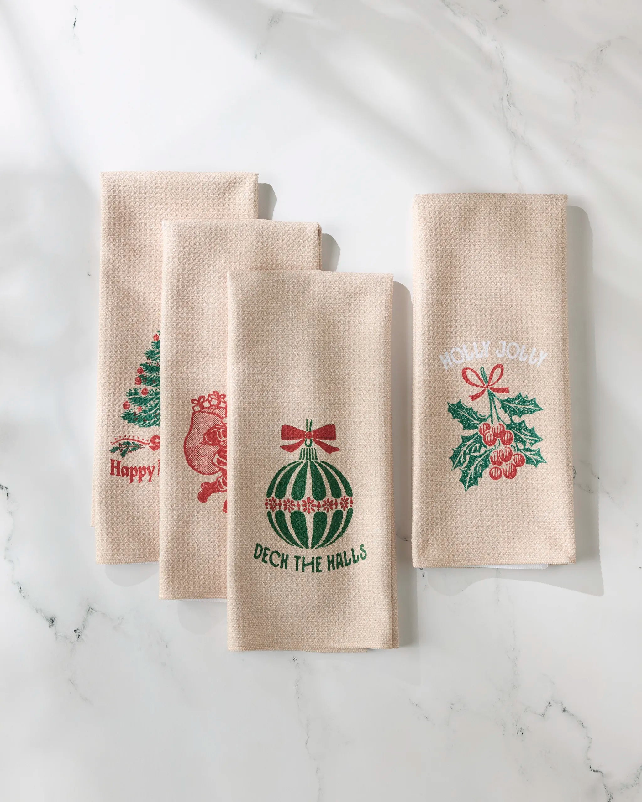 Four beige Festive Charm kitchen towels by GEOMETRY, each featuring a unique Christmas design and text: a tree, Santa, ornament, and holly, are displayed on a white surface.